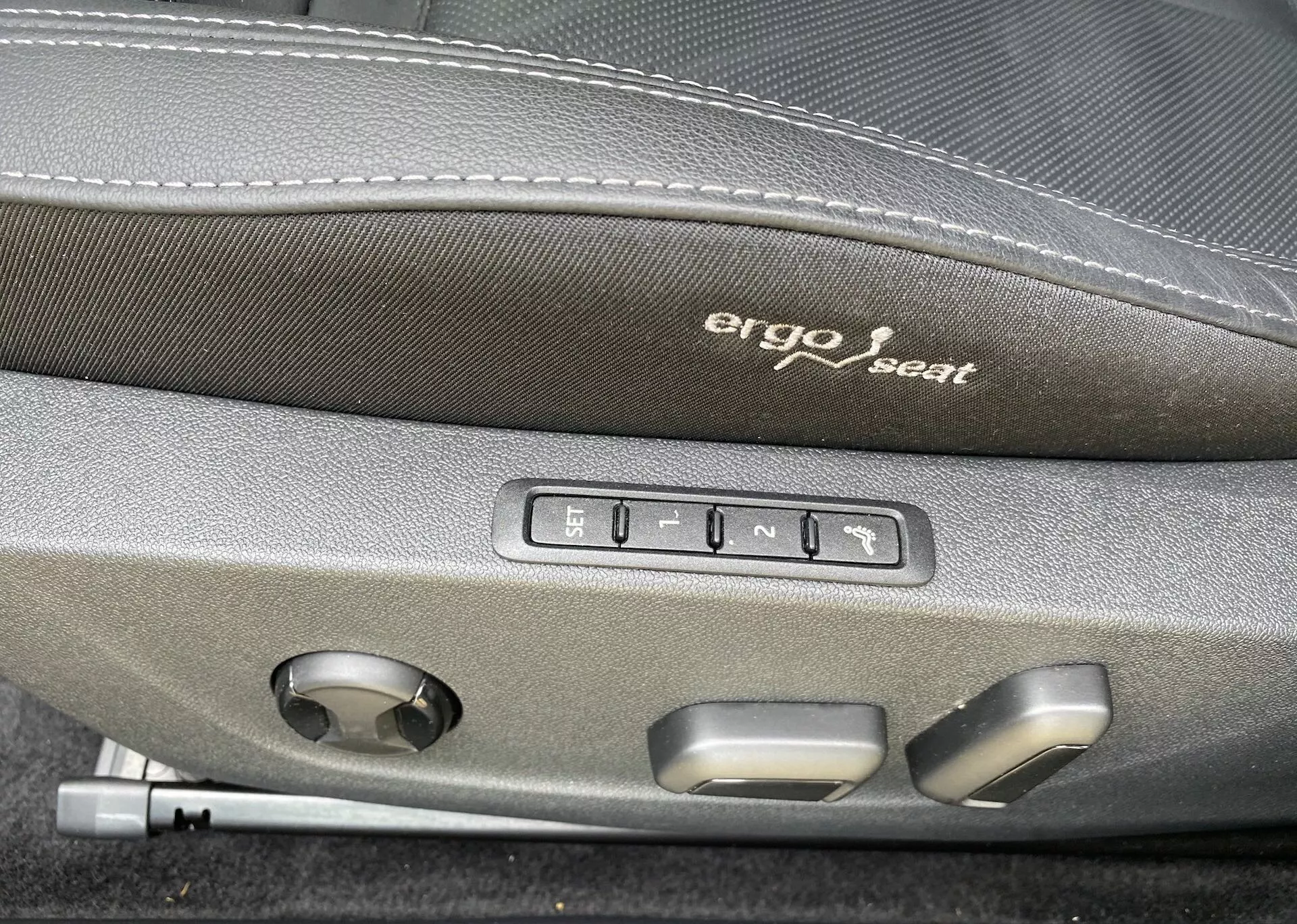 Närbild av interiören i en Škoda Octavia 2020, med fokus på de ergonomiska sätesjusteringskontrollerna. 'Ergo seat' är broderat på den mörka övre delen av sätet. Under broderiet syns fem knappar: en för minnessäten och fyra för sätesjusteringar som framåt, bakåt, uppåt, nedåt och ryggstödets lutning. Det finns också en rund ratt, förmodligen för att justera ländryggsstödet eller andra multifunktionella sätesjusteringar, vilket tyder på överlägsen komfort och avancerad teknik i detta fordon.