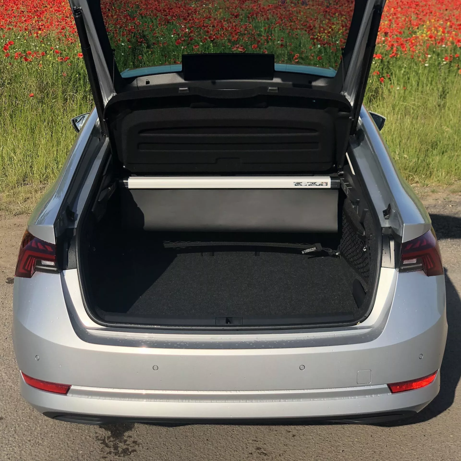 Vy från hög vinkel av en silverfärgad Skoda Octavia 2020 sedan med öppen bagagelucka, som visar det rymliga och organiserade lastutrymmet. Den bakre stötfångaren, de distinkta röda baklyktorna och en del av bilens emblem är synliga, med ett fält av röda blommor i bakgrunden.