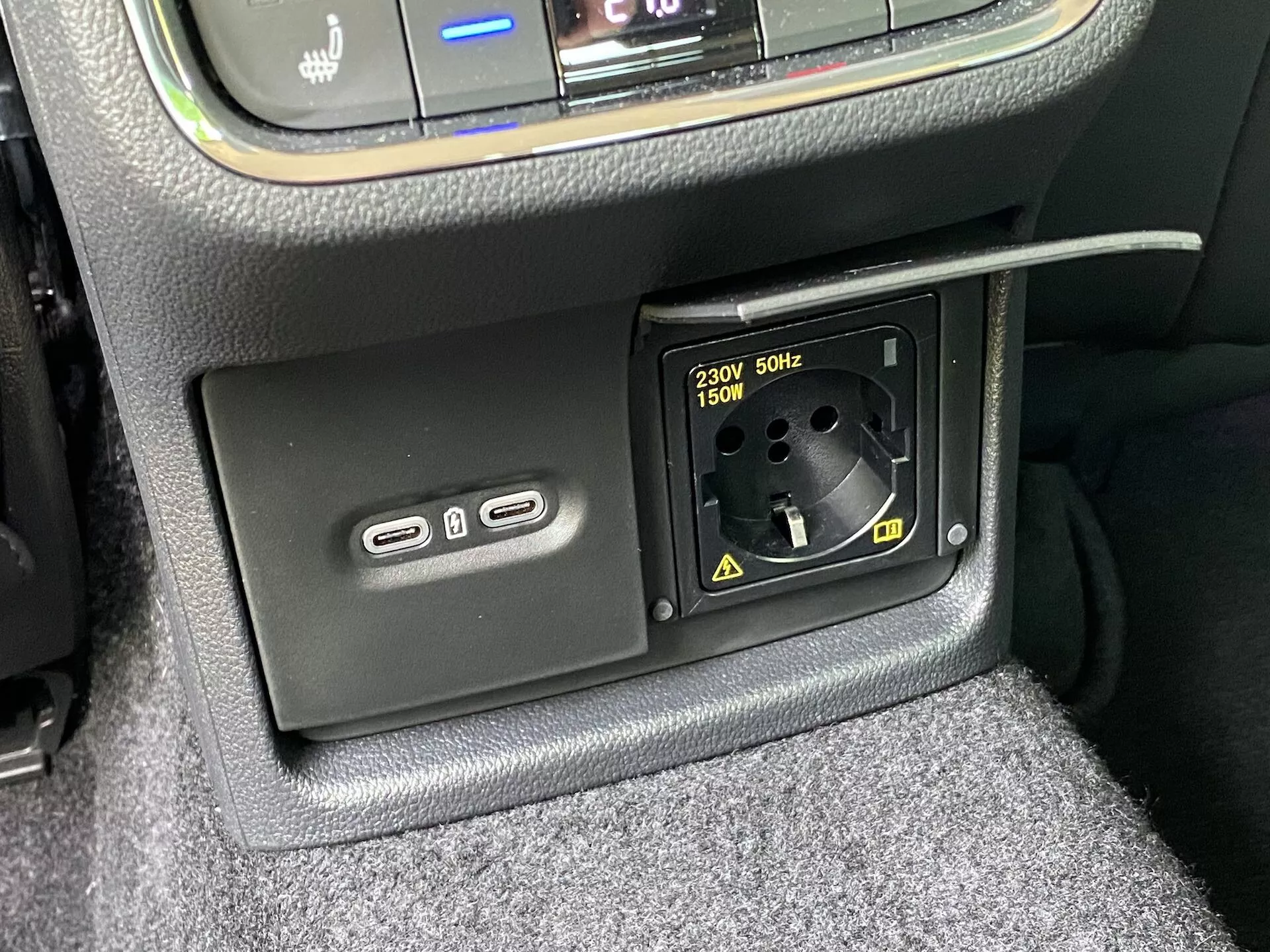 Närbild av Skoda Octavia 2020:s interiöra strömförsörjningsfunktioner som visar den nedre delen av mittkonsolen med två USB-A-portar för laddning eller multimedieanslutning till vänster och ett 230V AC-uttag för europeiska kontakter som stöder upp till 150W till höger, omgiven av texturerade ytor och synlig bilmatta.