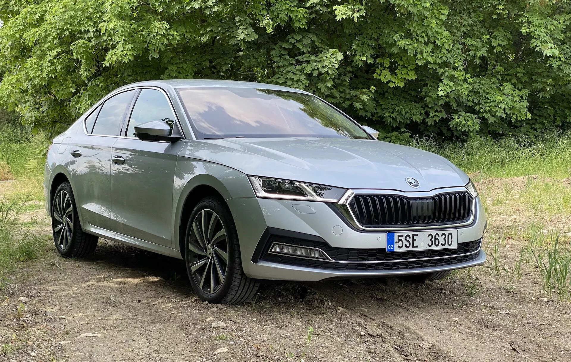 Trekvartsvy framifrån av en modern Skoda Octavia sedan från 2020, visad från en något lägre vinkel, med fokus på dess frontfasad, galler med vertikala lameller och en framträdande logotyp, eleganta horisontellt orienterade LED-strålkastare och undre stötfångare med dimljus och luftintag. Bilen är placerad på en icke-asfalterad yta med vegetation i bakgrunden, under naturligt dagsljus. Andra synliga detaljer inkluderar lättmetallfälgar med flerekrad design, en karaktärslinje som löper längs sidprofilen och dörrhandtag. Lämplig för att visa upp bil- och design och moderna fordonsfunktioner i en utomhusmiljö.