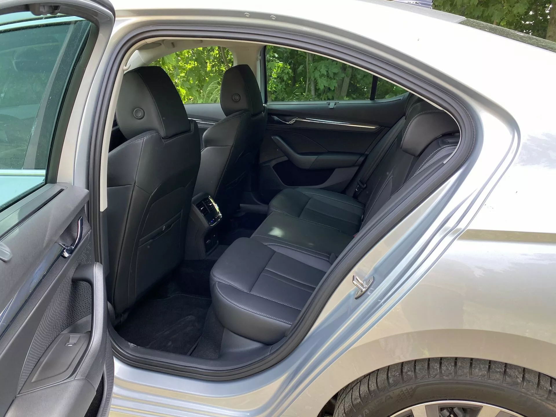 Bild av Skoda Octavia 2020:s interiör, med fokus på baksätet. Bakdörren är öppen och visar svarta lädersäten, nackstöd, säkerhetsbälten och ett uppfällt mittarmstöd. Dörrpanelen är tillverkad i ett läderliknande material för komfort. Synliga funktioner inkluderar förvaringsfickor på baksidan av framsätena, ren matta och ett fästpunkt för barnstol. Den omgivande belysningen tyder på dagsljus i en grön förortsområde.