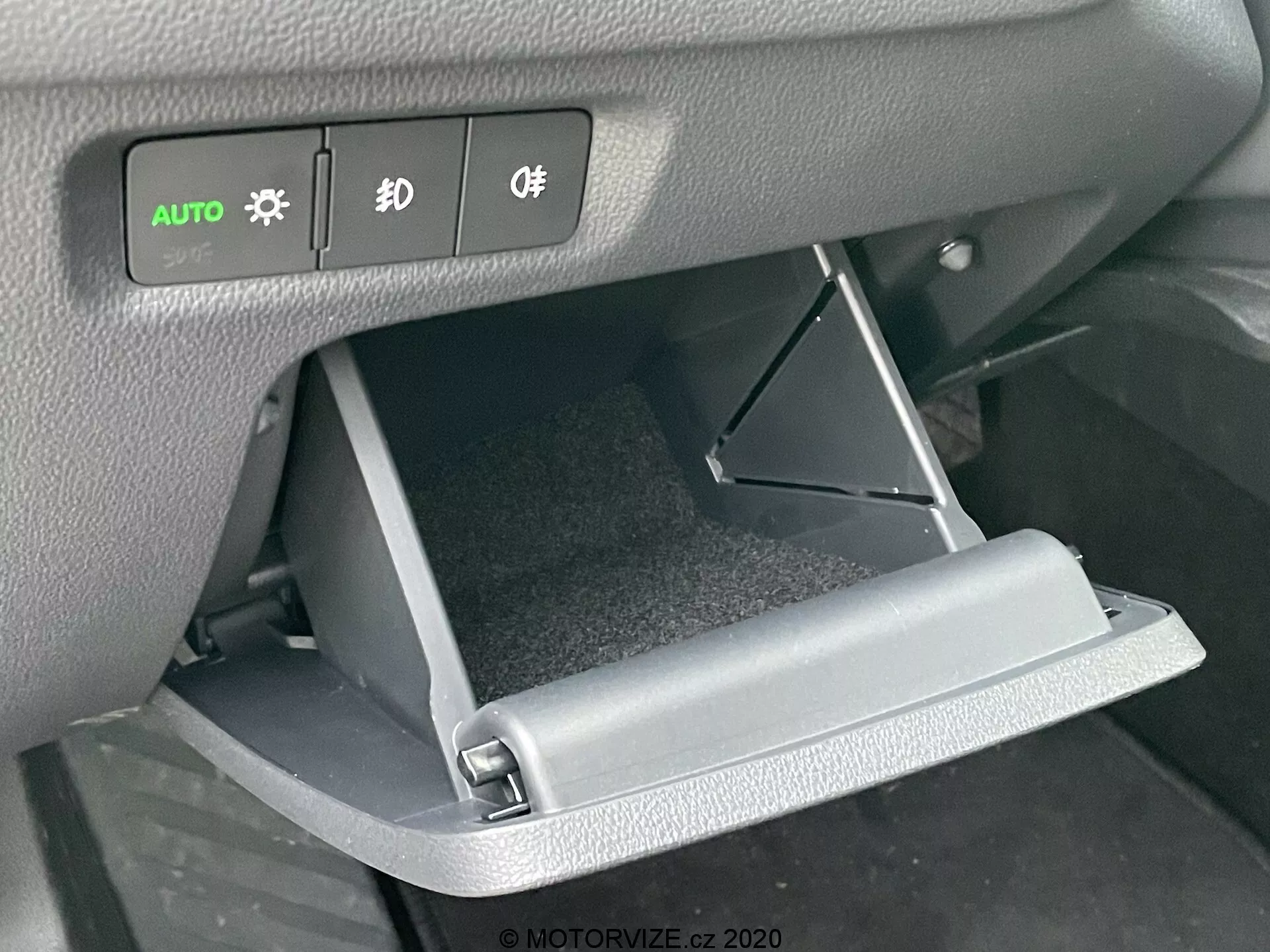 Närbild av interiören i Skoda Octavia 2020, med fokus på det öppnade handskfacket och instrumentpanelens reglage. Den främre vinkeln framhäver den nedåt öppnade handskfacksdörren och de upplysta knapparna för strålkastare, dimljus och belysningskontroller ovanför.