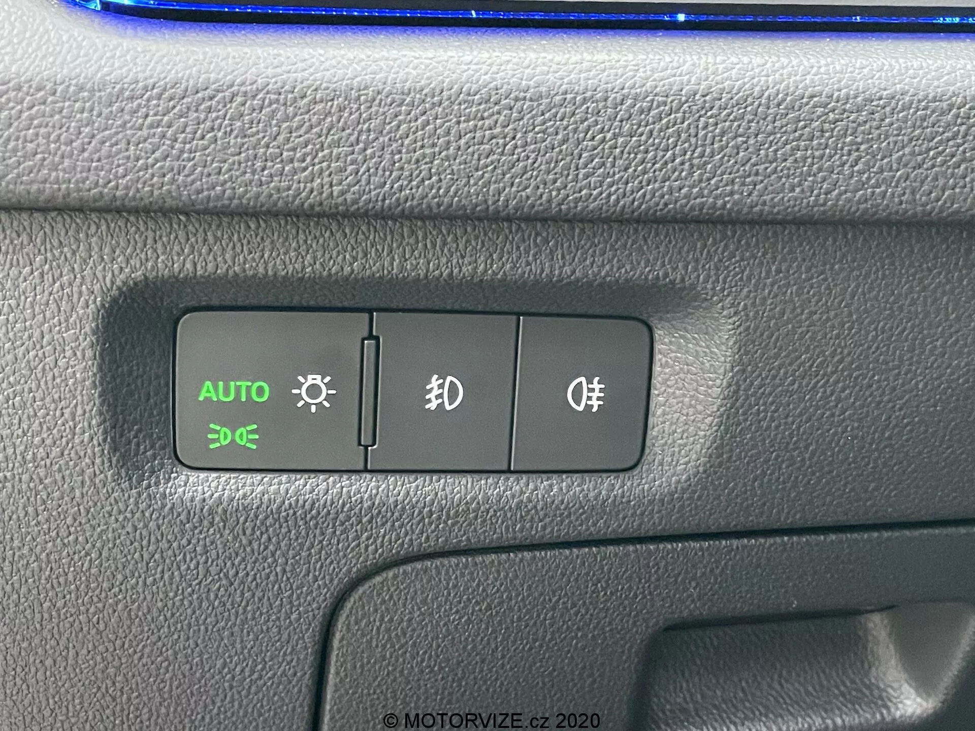 Närbild på en kontrollpanel i en Skoda Octavia 2020, med fokus på knapparna för automatiska strålkastare, parkeringsljus och dimljus. AUTO-knappen är upplyst i grönt, vilket indikerar att de automatiska strålkastarna är aktiva. Den texturerade plastpanelen har en tvåfärgad design med möjlig blå reflektion ovanför, i en bilinteriörinställning.