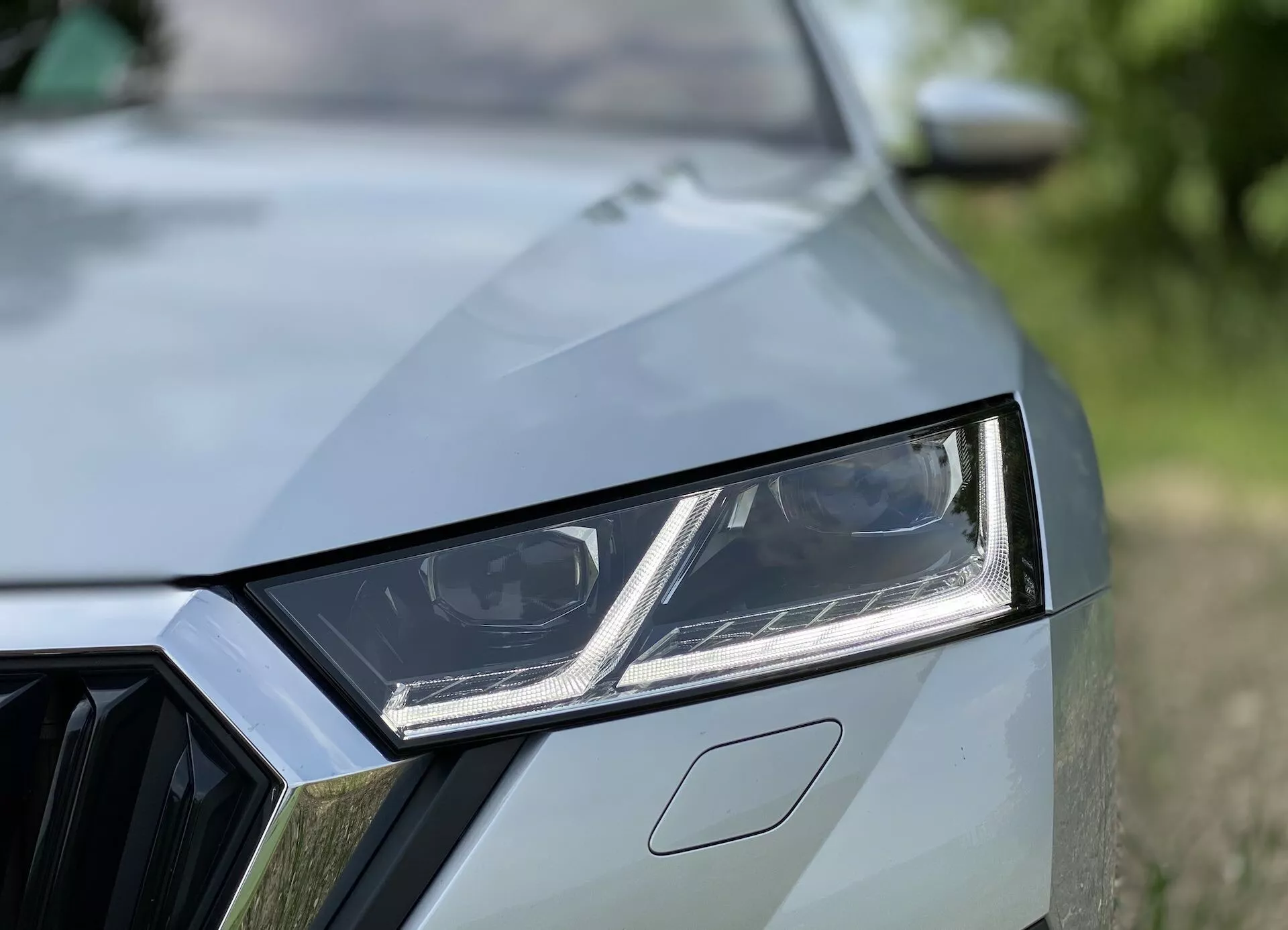 Närbild av den främre vänstra sidan av en modern bil som visar detaljerade designelement som den vinklade LED-strålkastaren med integrerade dagsljuslampor, en partiell vy av grillen med geometriskt mönster och motorhuven med distinkta veck. Den högkvalitativa lacken och det oskarpa bakgrunden betonar bilens sportiga och moderna estetik.