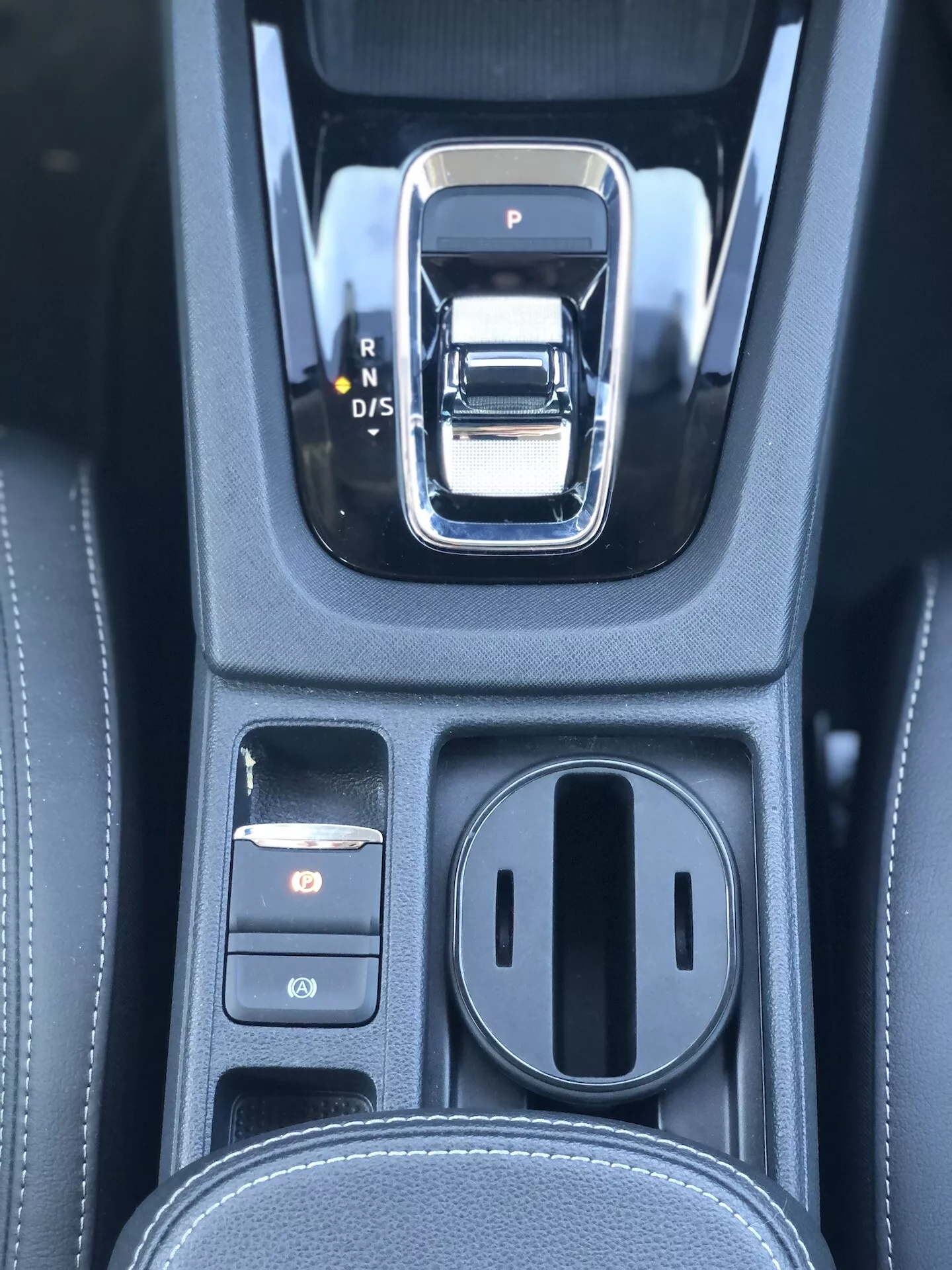 Närbild av området för automatisk växelspak i en 2020 Škoda Octavia Limousine. Växelspaken är en silver- och svart elektronisk spak med P, R, N och D/S alternativ. Till vänster finns två kontrollknappar, den övre lyser rött och är sannolikt den elektroniska parkeringsbromsen. Till höger finns ett täckt förvaringsfack med dubbla mugghållare. Den svarta konsolen har en läderliknande textur med detaljerade sömmar, vilket indikerar en högklassig interiör med avancerade transmissions- och elektroniska parkeringssystem.