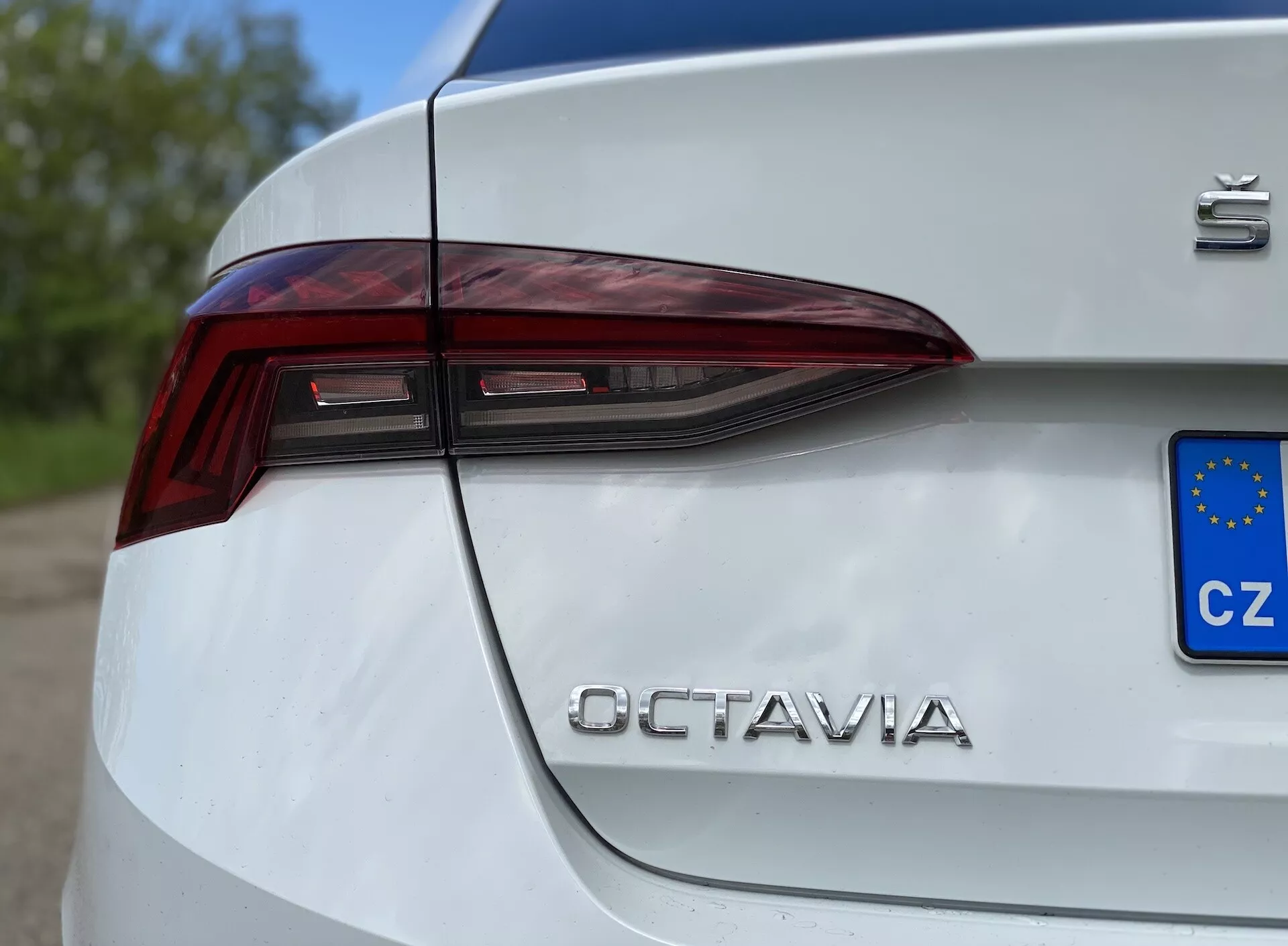 Närbild från sidan av bakre kvarten på en vit 2020 Škoda Octavia Limousine, som visar den skarpa, kantiga LED-bakljusdesignen som sträcker sig till bagageluckan, tillsammans med 'Škoda'-märket och krombokstäverna 'OCTAVIA', med en delvis synlig europeisk registreringsskylt (CZ), tagen från låg vinkel.