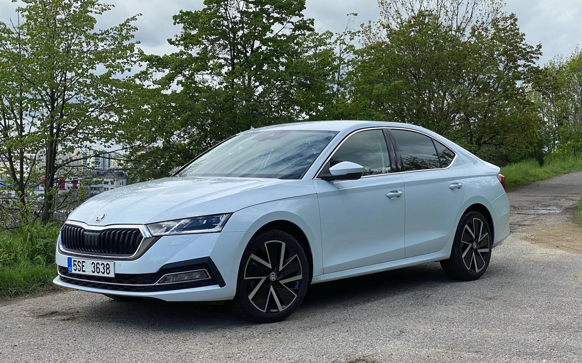 Måttligt vinklad sidovy av en ljusblå Skoda Octavia 2020 sedan, placerad vid vägkanten med grön grönska i bakgrunden. Visar en modern design med elegant fyrdörrars kaross, framträdande grill, aggressiva LED-strålkastare, stora flerspakslegeringshjul, karossfärgade sidobackspeglar med integrerade blinkers, sluttande taklinje, tonade fönster med kromkant, inbyggda dörrhandtag, låga däck och en badge som indikerar modell eller teknologipaket.