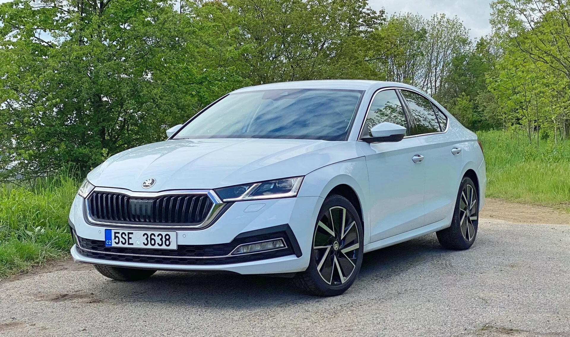 Bild av en ljusblågrå Škoda Octavia 2020 placerad på en asfalterad väg med lövverk i bakgrunden. Tagen i en trekvarts vinkel framifrån, framhäver fotot designens detaljer: den framträdande frontgrillen med vertikala ribbor och Škoda-emblemet, eleganta vinklade LED-strålkastare med dagsljus, separata dimljus, stora multiekrade aluminiumfälgar, sidobackspeglar med integrerade blinkers, potentiella främre parkeringssensorer och dynamiska karosslinjer längs huven och sidorna. Scenen antyder en lugn, dagsmiljö med naturligt ljus.