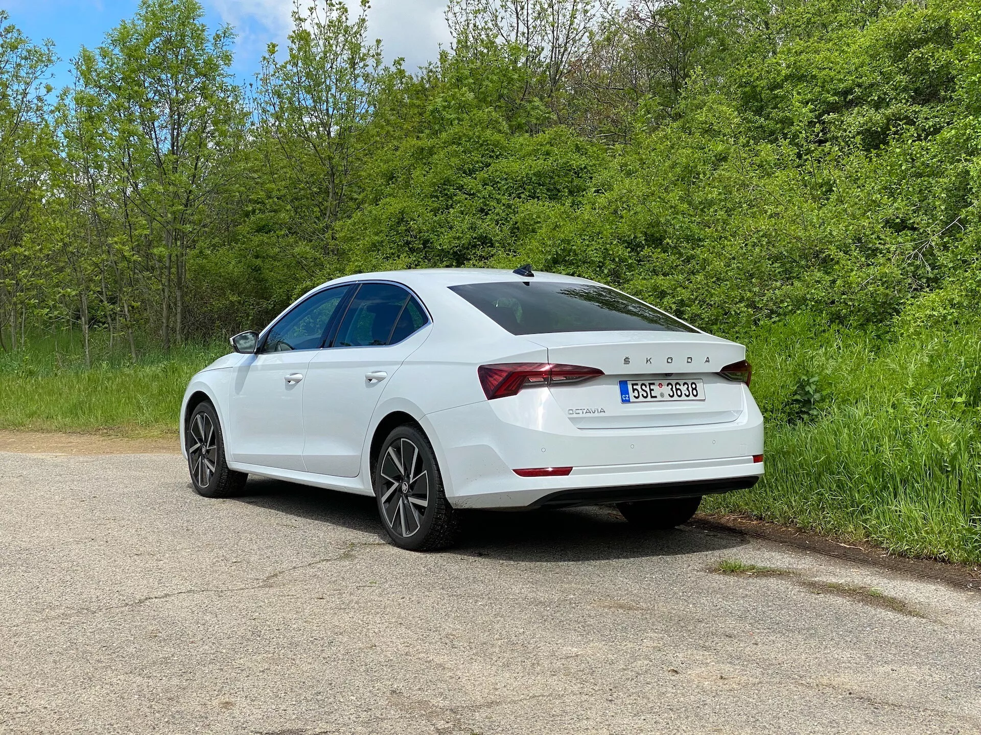 Vit sedan Skoda Octavia 2020 i trekvart bakrevy vinkel parkerad på en grusväg, flankerad av grönska, med detaljerad vy över karakteristiska baklyktor, bakspoiler, lättmetallfälgar och ett synligt soltak, som betonar dess moderna design och tekniska funktioner inklusive parkeringssensor på stötfångaren.