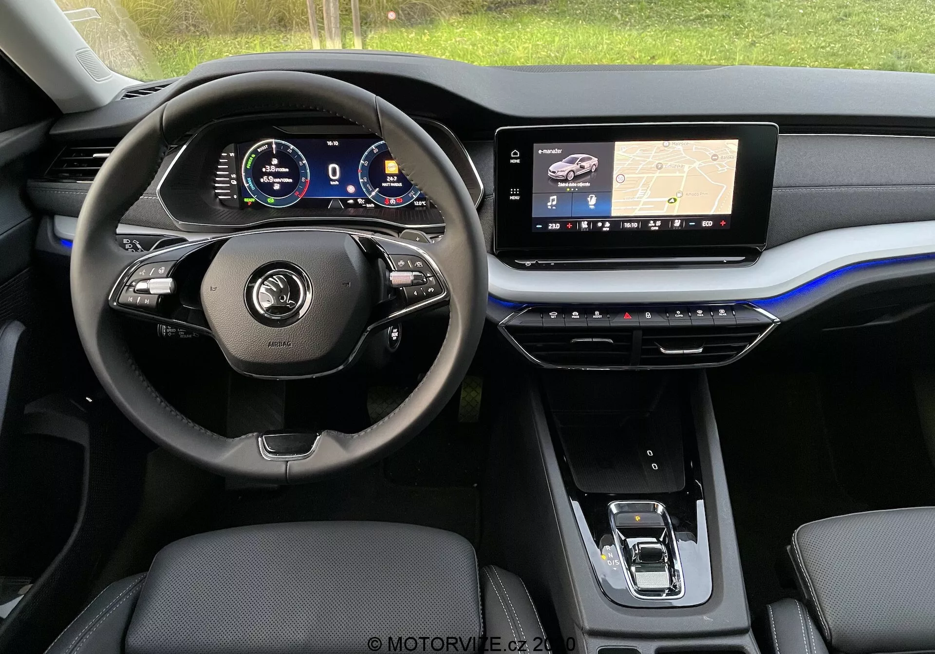 Interiörvy av en modern Skoda Octavia Limousine 2020 från baksätet, som visar instrumentbrädan och fokuserar på förarplatsen. Ratten har multifunktionsknappar och läderklädsel, bakom finns en digital instrumentpanel som visar olika fordonsmätare. Den centrala instrumentbrädan innehåller en stor infotainmentskärm som visar en navigationskarta med GPS-teknik, medan mittkonsolen nedanför innehåller tryckkänsliga knappar för klimatkontroll. Synliga är framsäten klädda i läderliknande material och en elektronisk växelväljare. Estetiken är elegant med mörka toner, metalliska accenter och en vågad blå highlight längs instrumentbrädan.
