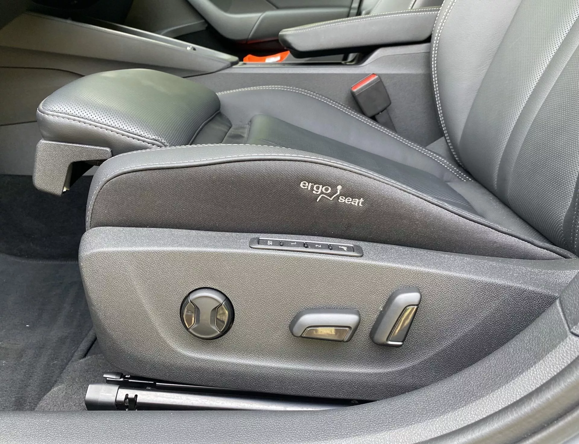 Närbild av Skoda Octavia 2020 förarsidokontroller som visar ergonomisk design med mörkt tygklädsel, en synlig märkning 'ergo seat' och elektroniska justeringskontroller inklusive en cirkulär lutningsjusterare och två metalliska positioneringsknappar, vilket lyfter fram fordonets premium- och teknologiskt avancerade interiör.