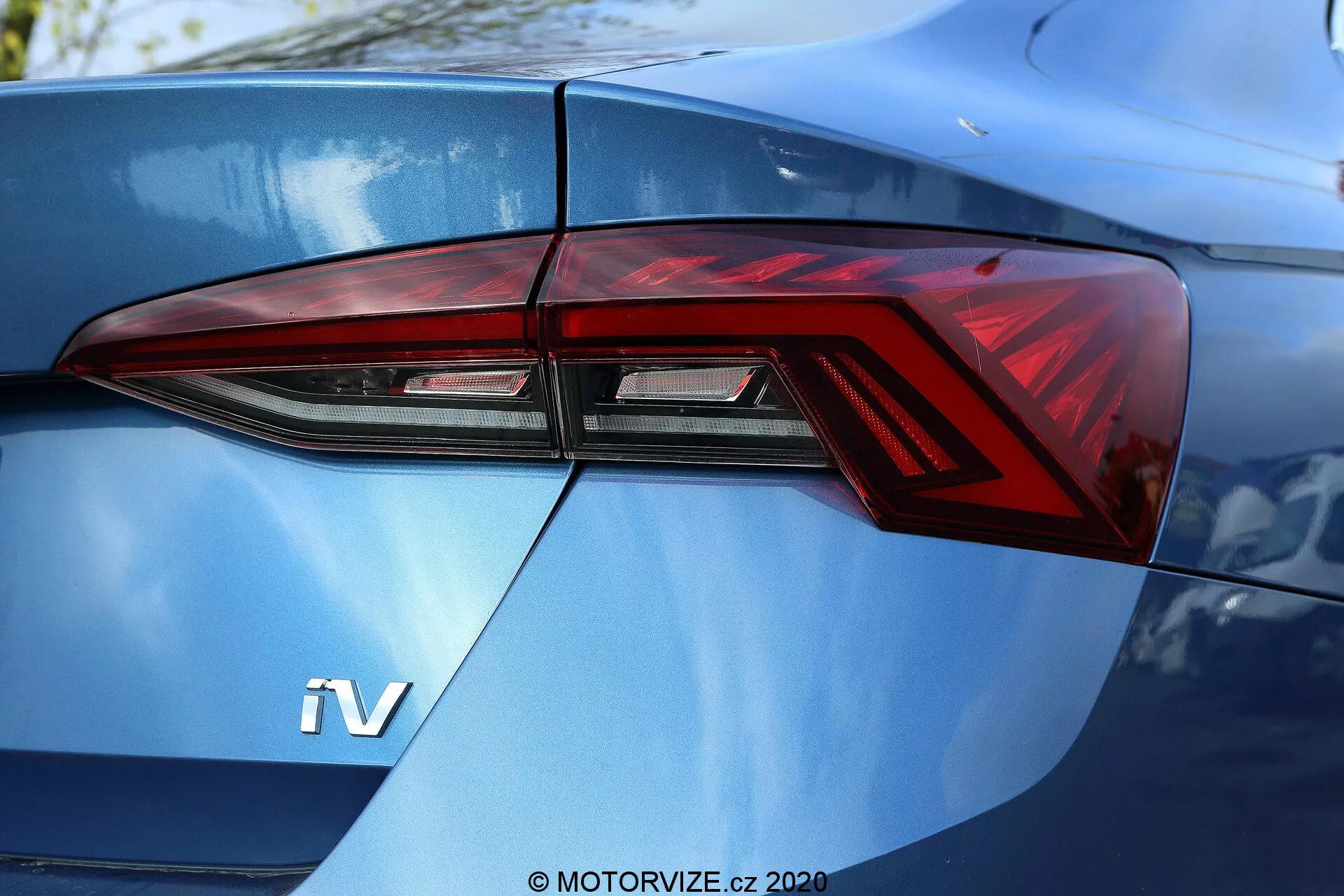 Närbild av bakdelen på en 2020 Skoda Octavia Limousine, med fokus främst på vänster baklykta sedd ur trekvarts vinkel. Den moderna baklyktedesignen har skarpa vinklar och distinkta LED-lyselement som är sömlöst integrerade i bilens karosslinjer. En synlig 'iV'-symbol på den glänsande metallic blåa bakluckan antyder att fordonet är en hybrid eller elbil.