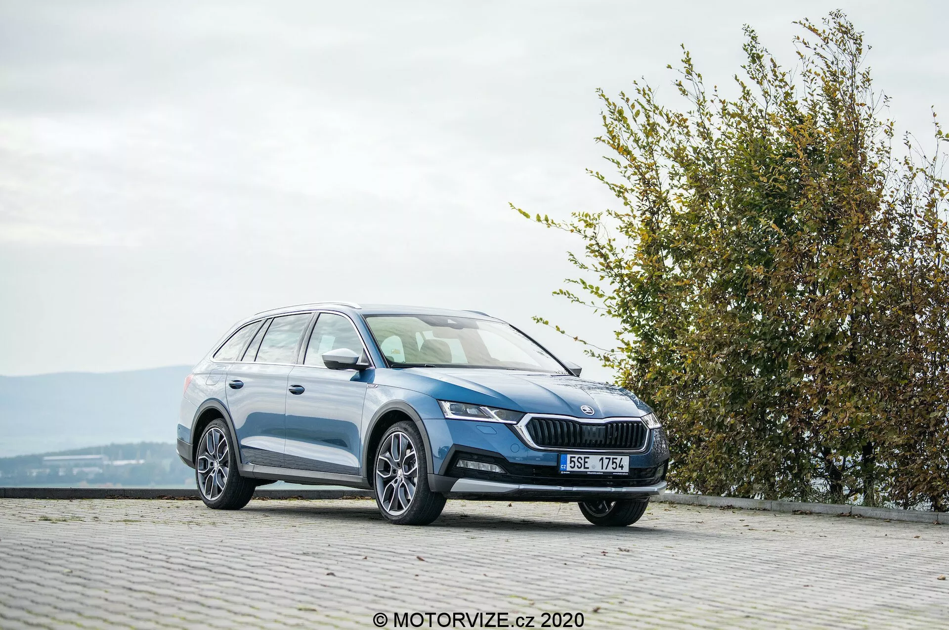 Fullstor kombi Skoda Octavia Scout 2020, fotograferad från en trekvarts framåtvinkel på ett stenbelagt område på en upphöjd plats. Bilden visar frontgrillen med en framträdande emblem, eleganta, vinklade strålkastare, integrerade dimljus, slät sidoprofil med lättmetallfälgar, takräcken och en blank färgfinish. Den omgivande kontexten inkluderar en häck eller buske till höger, vilket tyder på närhet till en park eller grönområde, under gynnsamma väderförhållanden.