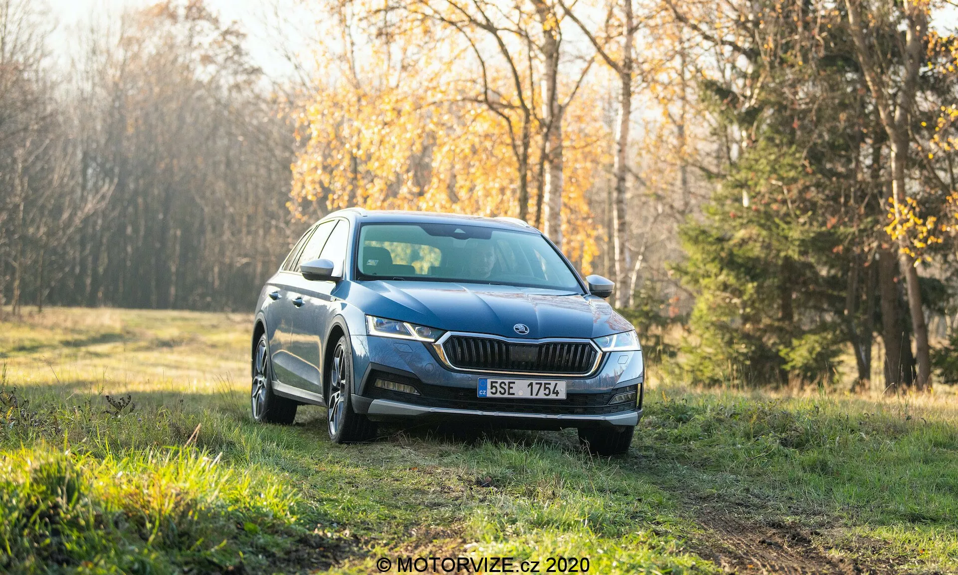 En blå Skoda Octavia Scout 2020 sedan, fotograferad från en trekvarts vinkel framifrån, parkerad på ett gräsbevuxet område med en skog i bakgrunden. Bilden visar moderna LED-strålkastare, ett distinkt främre galler, dimljus, flerspokriga fälgar och integrerade blinkers i sidospeglarna. Miljön verkar vara tidig morgon eller sen eftermiddag med mjukt solljus som ger en varm ton till den rena bilen avsedd för reklam eller översikt. Den suddiga bakgrunden framhäver bilens designegenskaper och attraktiva utomhusmiljö.