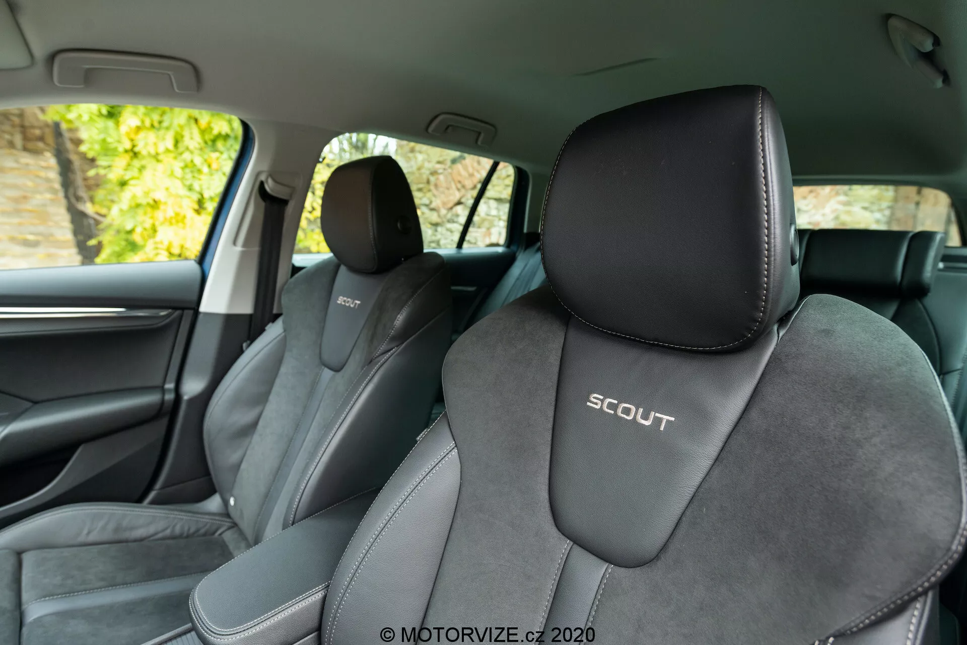Interiörvy av en Skoda Octavia Scout 2020 från baksätet, som visar framsäten. Vinkeln gynnar den vänstra sidan och ger en klar vy av förarsätet med en präglad 'SCOUT'-logotyp. Sätena är klädda i tvåfärgade toner, övervägande svarta med ljusare grå inslag och en mittdel av mockaliknande material, som framhäver en sportig design. Bilden visar också B-stolpen, mörka innertillbehör, en del av framdörren, ett högtalargaller och ett kromat dörrhandtag. Bakgrunden utanför fönstren verkar visa lövverk, vilket tyder på att bilen är parkerad utomhus i naturligt ljus.