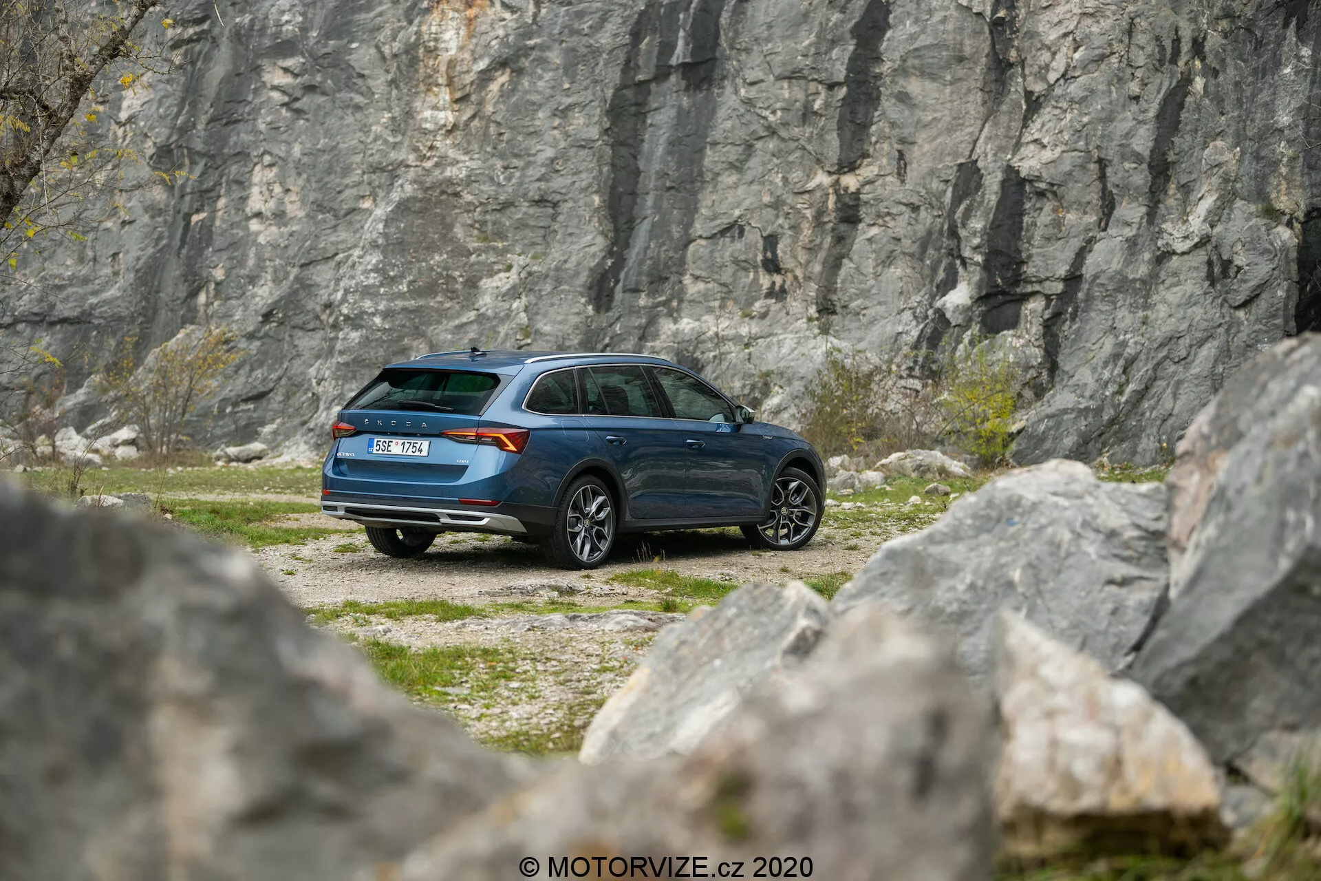 Bilden visar en Skoda Octavia Scout 2020 parkerad i en stenig terräng, med ett stenbrott eller en klippvägg i bakgrunden, fångad från ett lätt teleobjektivsperspektiv. Den trekvarts bakre vinkeln ger en detaljerad vy av fordonets sidoprofil och bakre ände, inklusive det moderna kombidesignen, sluttande bakre taklinjen och den mörkblå lacken. Synliga funktioner inkluderar bakljusmonteringen, fönstrens form, bakrutetorkaren, hjuldesignen och sidospeglarna. Miljön antyder teman av äventyr och utforskning, vilket indikerar fordonets lämplighet för avlägsna eller utmanande terränger.