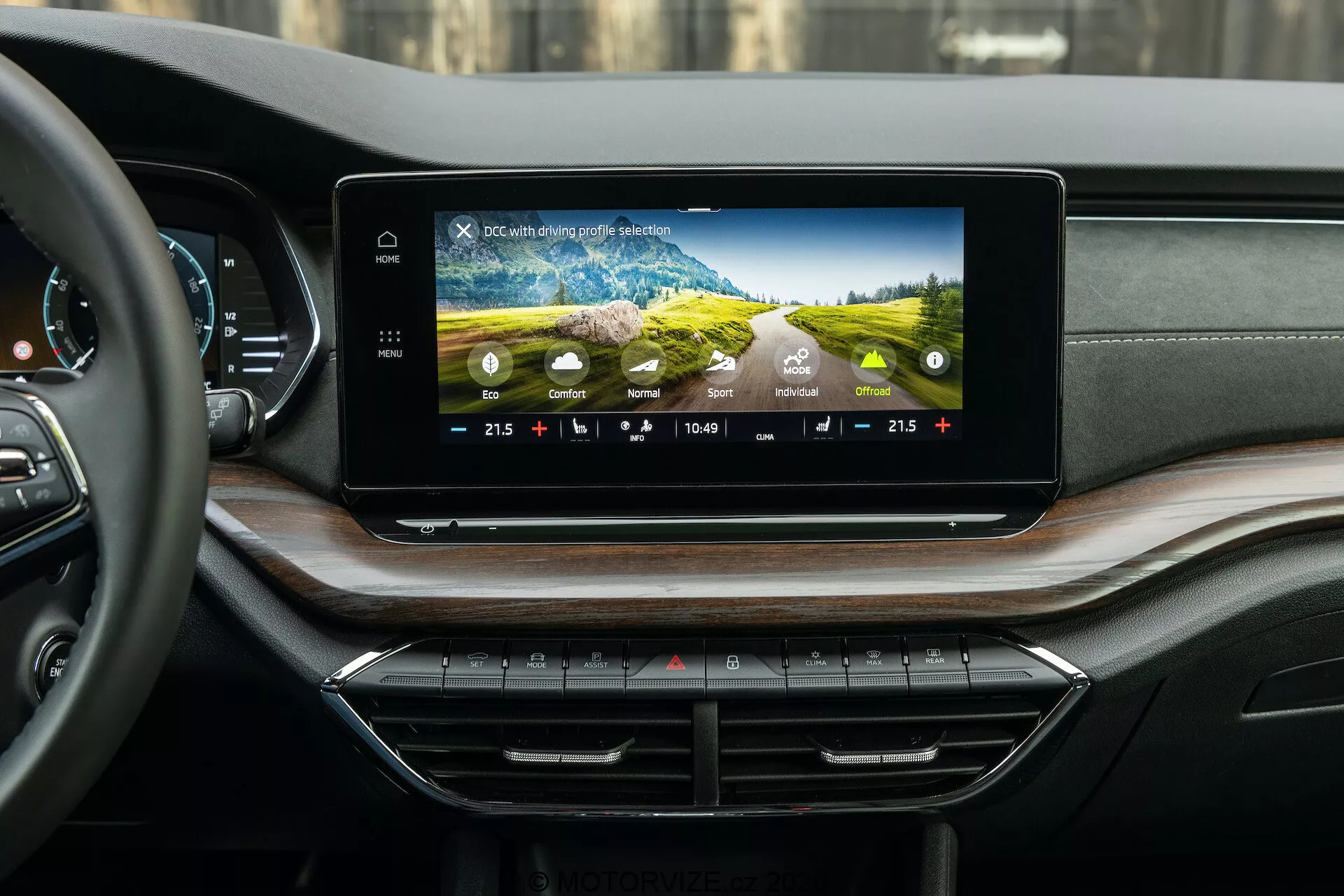 Interiörvy av Skoda Octavia 2020 (Octavia Scout) som visar det moderna infotainmentsystemet och instrumentbrädan, fotograferat från bakpassagerarens perspektiv. Den breda, horisontellt orienterade pekskärmen på instrumentbrädan visar färggrafik, klimatkontrollinställningar, tid och utomhustemperatur. Nedanför finns snyggt ordnade klimatkontrollknappar och rattar. Den lyxiga instrumentbrädan har material med mjuk känsla och träfaner, med diskret integrerade luftventiler.