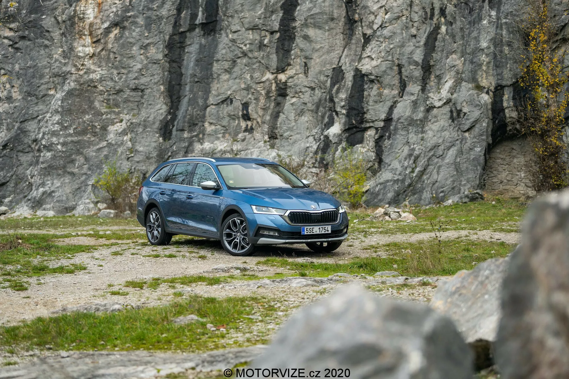 Sid-framvy av en blå Skoda Octavia Scout 2020 kombi på grusigt terräng med en bergig bakgrund, som visar moderna LED-strålkastare, framträdande grill, lättmetallfälgar, sidospeglar, fönsterlister, takräcken och en ren metallisk finish, vilket antyder avancerade funktioner som belysningsteknik, däcktrycksövervakning och möjliga säkerhetssystem.