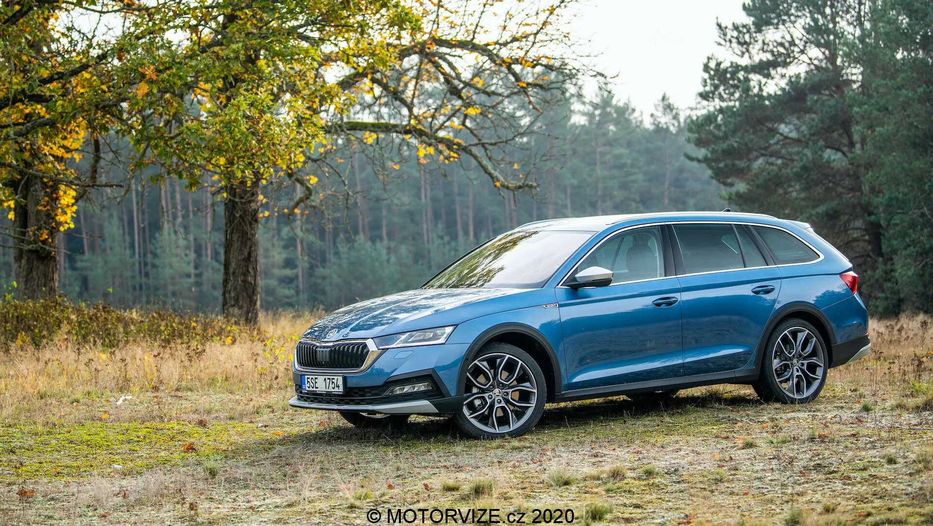 Sidoprofil av en blå Skoda Octavia Scout 2020 kombi på en grusväg med träd och gräs i bakgrunden, som visar dess förmåga att hantera terrängkörning. Bilen har en metallic lack, mångekrade fälgar, LED-strålkastare, ett distinkt främre galler, takräcken, karossfärgade sidobackspeglar och dörrhandtag samt ett soltak. Designfunktionerna inkluderar en karaktärslinje som går från främre hjulbågen till bakljusen och omslutande bakljus.