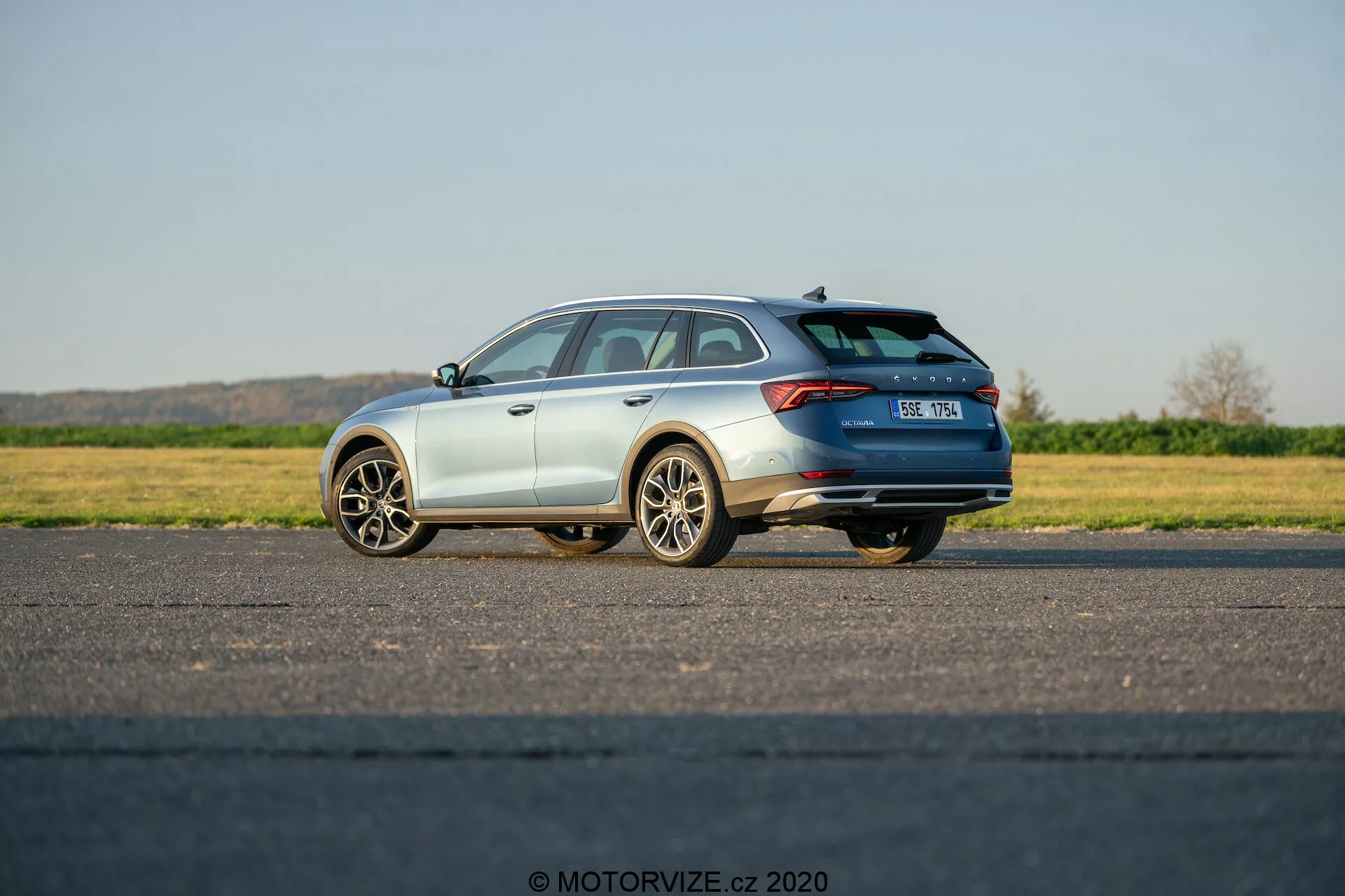 Bilden visar en Škoda Octavia Scout 2020, en modern kombibil, fångad med en trekvarts bakre vy. Den framhäver bilens designinslag inklusive de horisontella bakljusen som sträcker sig mot centrum av bakluckan, takspoilern, takräcken och flerspakiga lättmetallfälgar. Bilen står på en platt asfalterad yta med förarsidan närmare kameran. Bilen har en metallic lackering och en del av avgassystemet är synligt. Registreringsskylten är något suddig och Škoda-emblemet är synligt. Fotograferad i naturligt dagsljus under klar himmel, visar bilden en stillastående bil med potentiellt dolda tekniska funktioner som parkeringshjälp, backkameror eller kollisionsundvikande system.