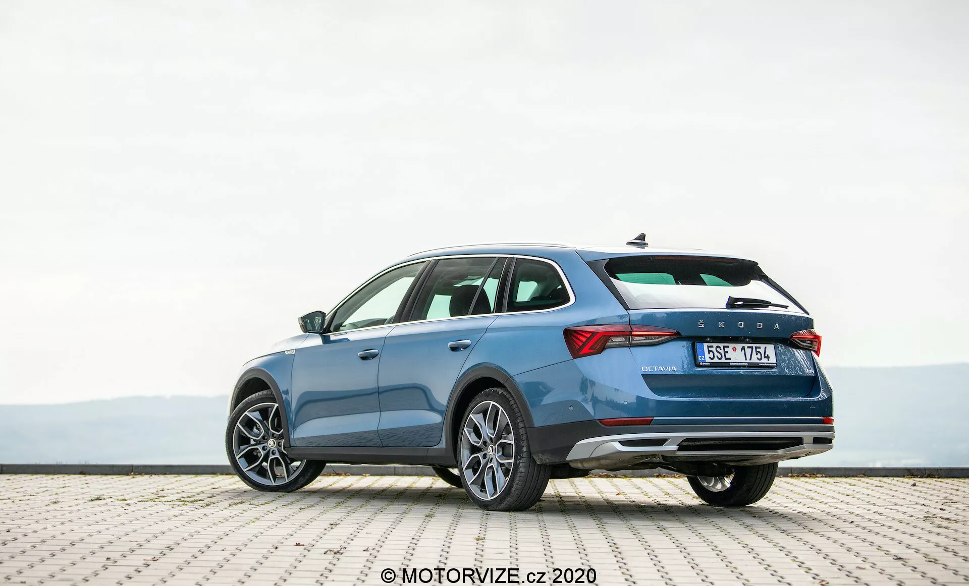En detaljerad bild av en blå Skoda Octavia Scout kombi från 2020, parkerad på en kaklad yta, tagen från ett bakre trekvartsperspektiv. Fotografin framhäver fordonets eleganta profil, distinkta bakluckedesign och högra sida. De synliga funktionerna inkluderar horisontella baklyktor, en karaktärslinje som antyder rörelse, en bakre stötfångare med reflektorer som möjligen innehåller parkeringssensorer, dubbla avgasrör och flerspakiga aluminiumfälgar. Bilden antyder en rymlig interiör, avancerade säkerhetsfunktioner och samtida designelement som är lämpliga för entusiaster av bilteknik.