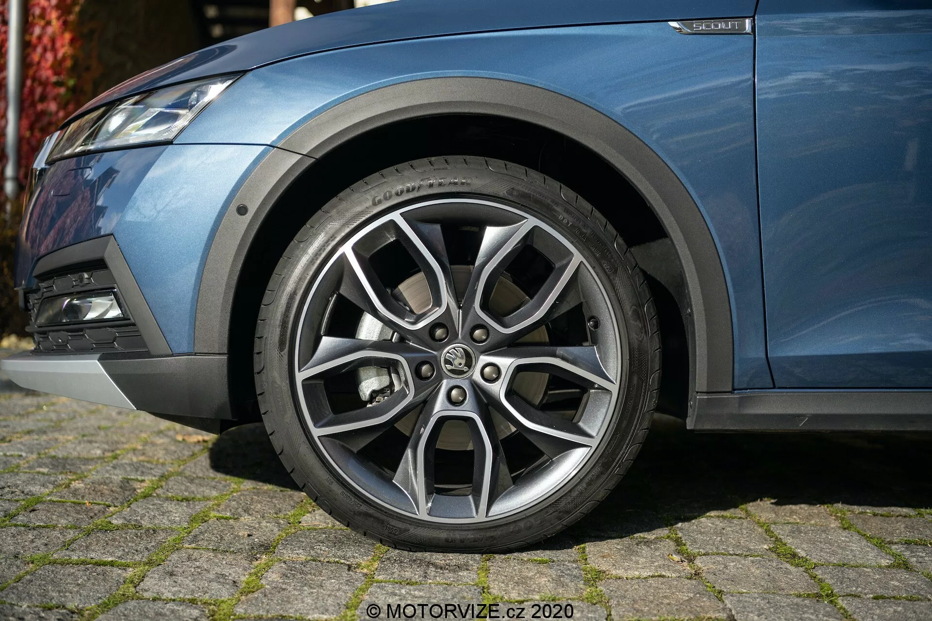 Närbild av vänster framsida av en Skoda Octavia 2020 (Octavia-Scout) med fokus på det noggrant designade lättmetallhjulet med flerspoksdesign, kontrasterande silverfinish och synliga mönster i slitbanan. Fotot, taget från en låg vinkel, fångar en del av bilens metalliska blå kaross, främre stötfångare med dimljus och fjädringskomponenter, som visar upp dess moderna, sportiga utseende.
