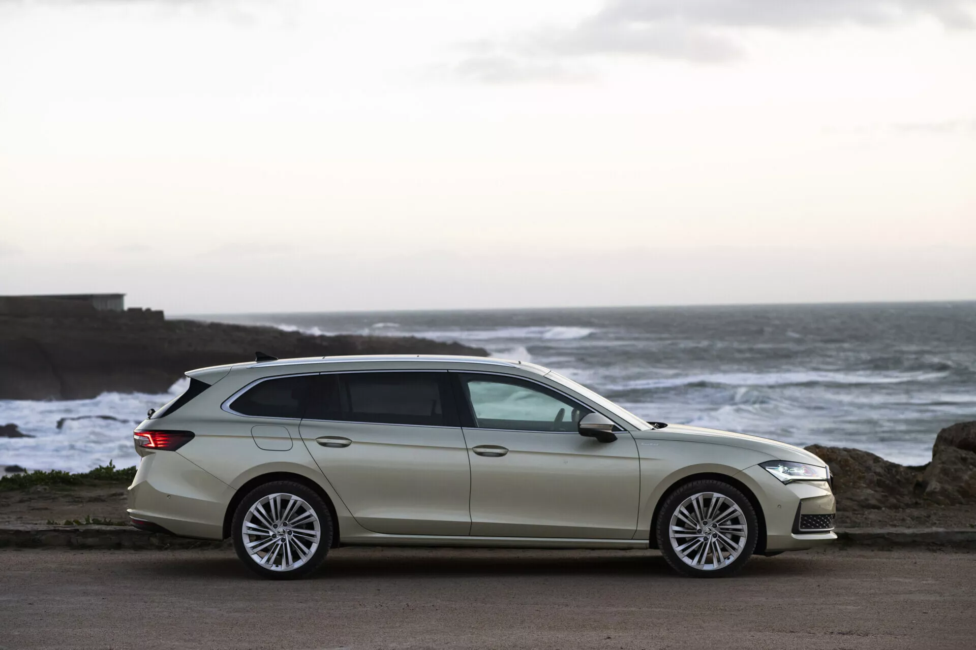 Skoda Superb Combi 2024 i ljus nyans parkerad nära kusten i skymningen, framhäver dess moderna design, strömlinjeformade kaross och premium flerspakiga legeringshjul