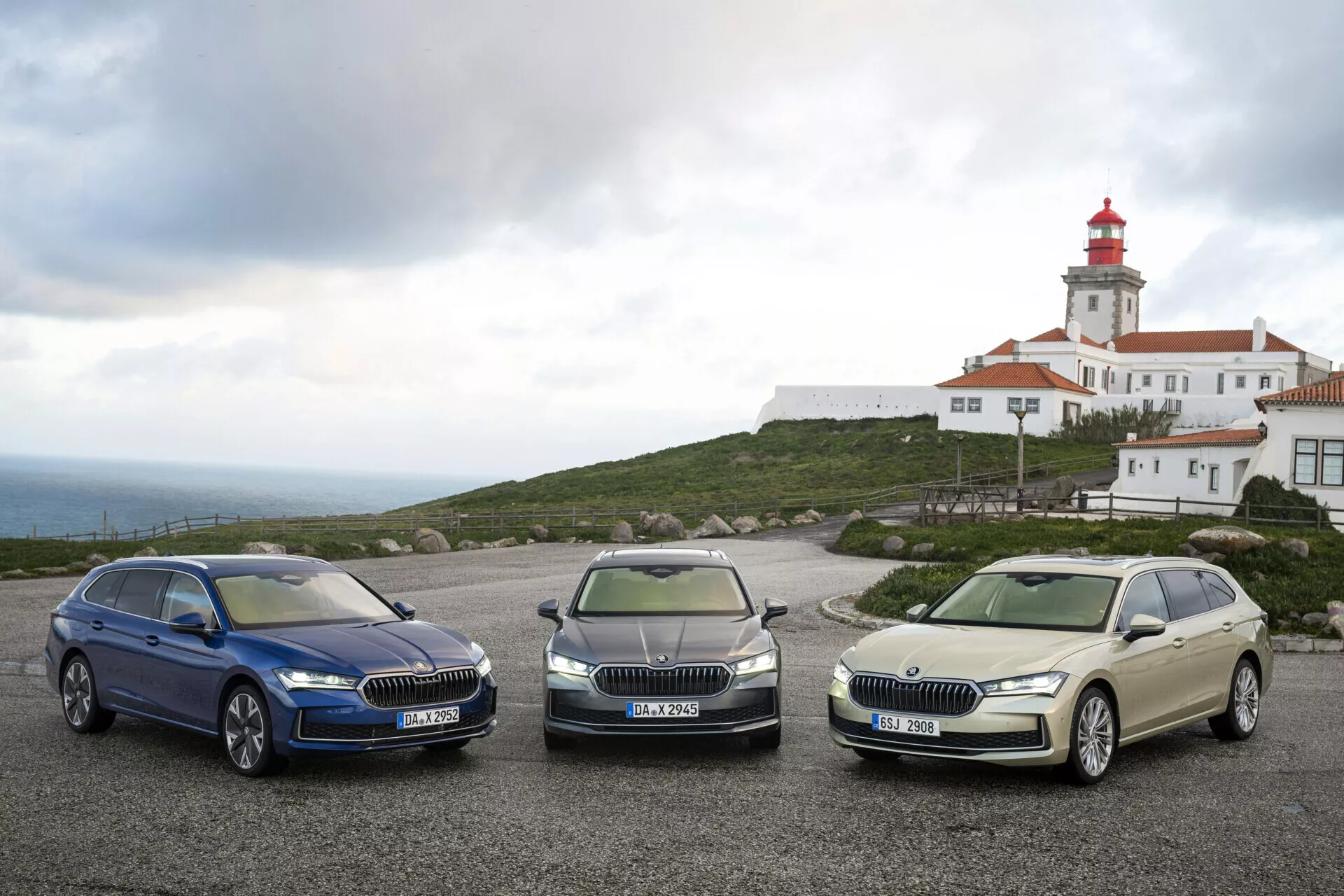 2024 års Skoda Superb Combi-modeller i mörkblått, metallgrått och blekguld parkerade bredvid varandra som uppvisar LED-strålkastare, kromgrillar och lättmetallfälgar med ett fyrtorn och en byggnadsstruktur i bakgrunden under en molnig himmel