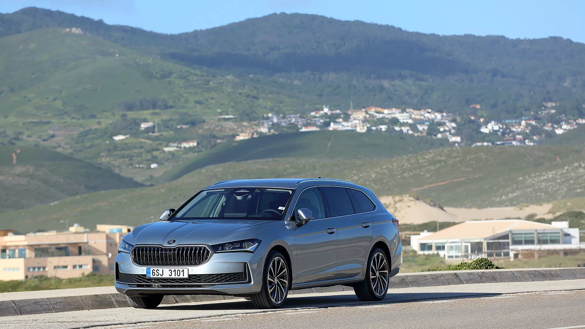 Skoda Superb Combi 2024 i en tredjedels framsida vy på en väg med en kuperad landskapsbakgrund, med en framträdande grill, LED-strålkastare och panoramatak