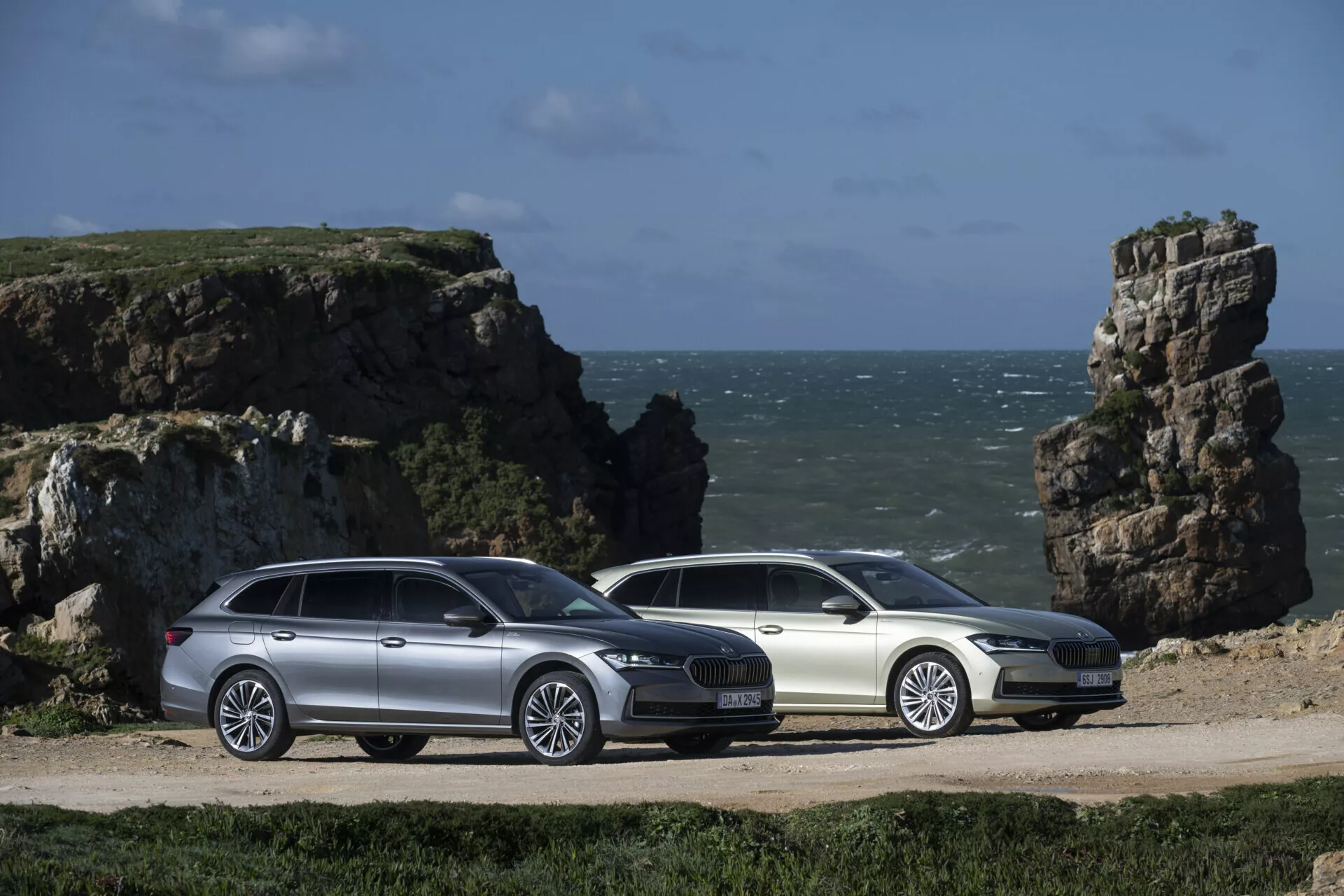 2024 Skoda Superb Combi stationsvagnar i silver och vitt, visas sida vid sida i en tre kvarts vy som visar deras aerodynamiska profiler, legeringshjul och integrerade takräcken, inställda mot en kustbakgrund med klippiga utskott.