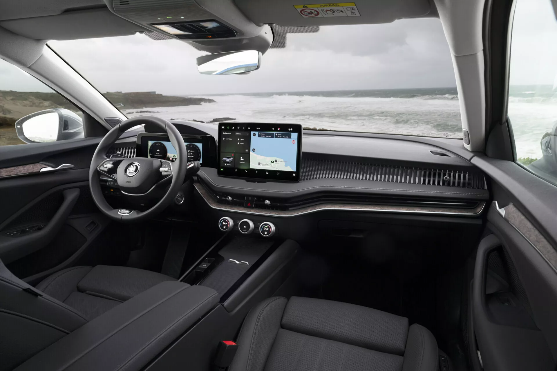 Interiör i Skoda Superb Combi 2024 som visar instrumentbrädan från baksätets perspektiv, med fokus på multifunktionsratten, horisontellt pekskärmsinfotainmentsystem, dekorativ instrumentbrädastrimma, mörka lädersäten och detaljer om panoramatak.