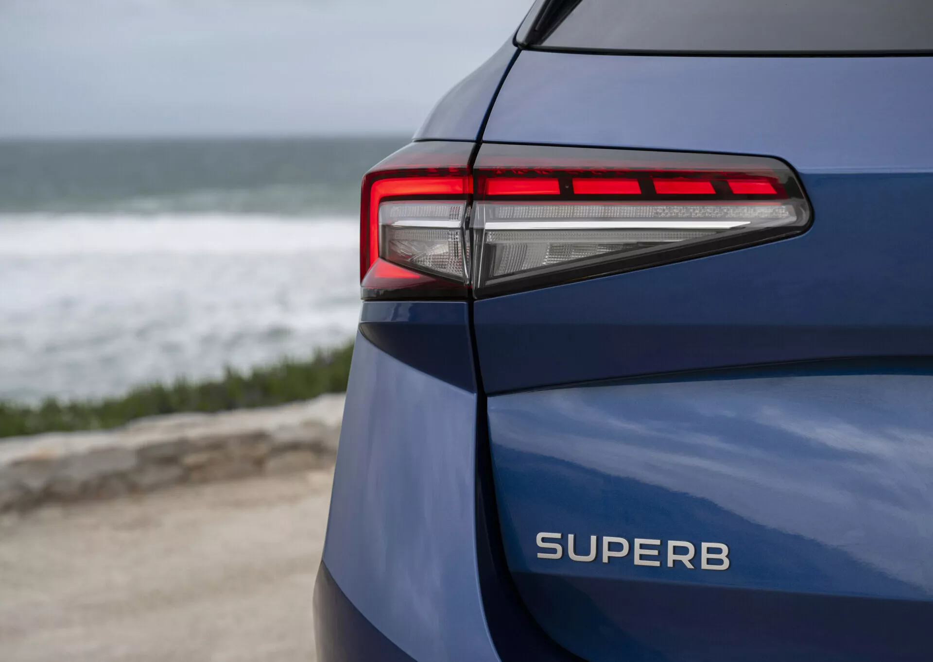 Škoda Superb Combi 2024 i blått, visar dess bakre vänstra sida, tydligt visar modellnamnet SUPERB, LED-bakljus med moderna horisontella linjer och skarpa vinklar mot en kustbakgrund