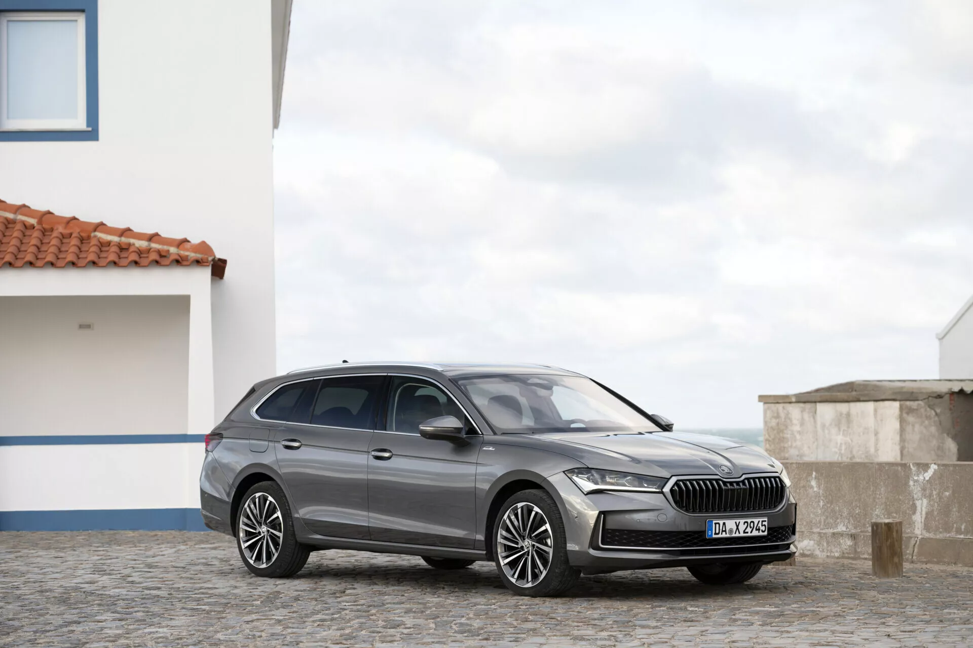 Skoda Superb Combi 2024 fångad från ett tre fjärdedels frontvinkel som lyfter fram dess kromaccentuerade grill, LED-strålkastare, aerodynamiska huvs konturer, flerspakiga lättmetallhjul, karossfärgade sidospeglar med möjliga integrerade blinkers, i en metallisk finish med lätt tonade fönster och integritetsglas, som visar den lyxiga stationvagnens design och funktioner på ett kullerstensbelagt underlag med klar himmel och bostadsbakgrund.