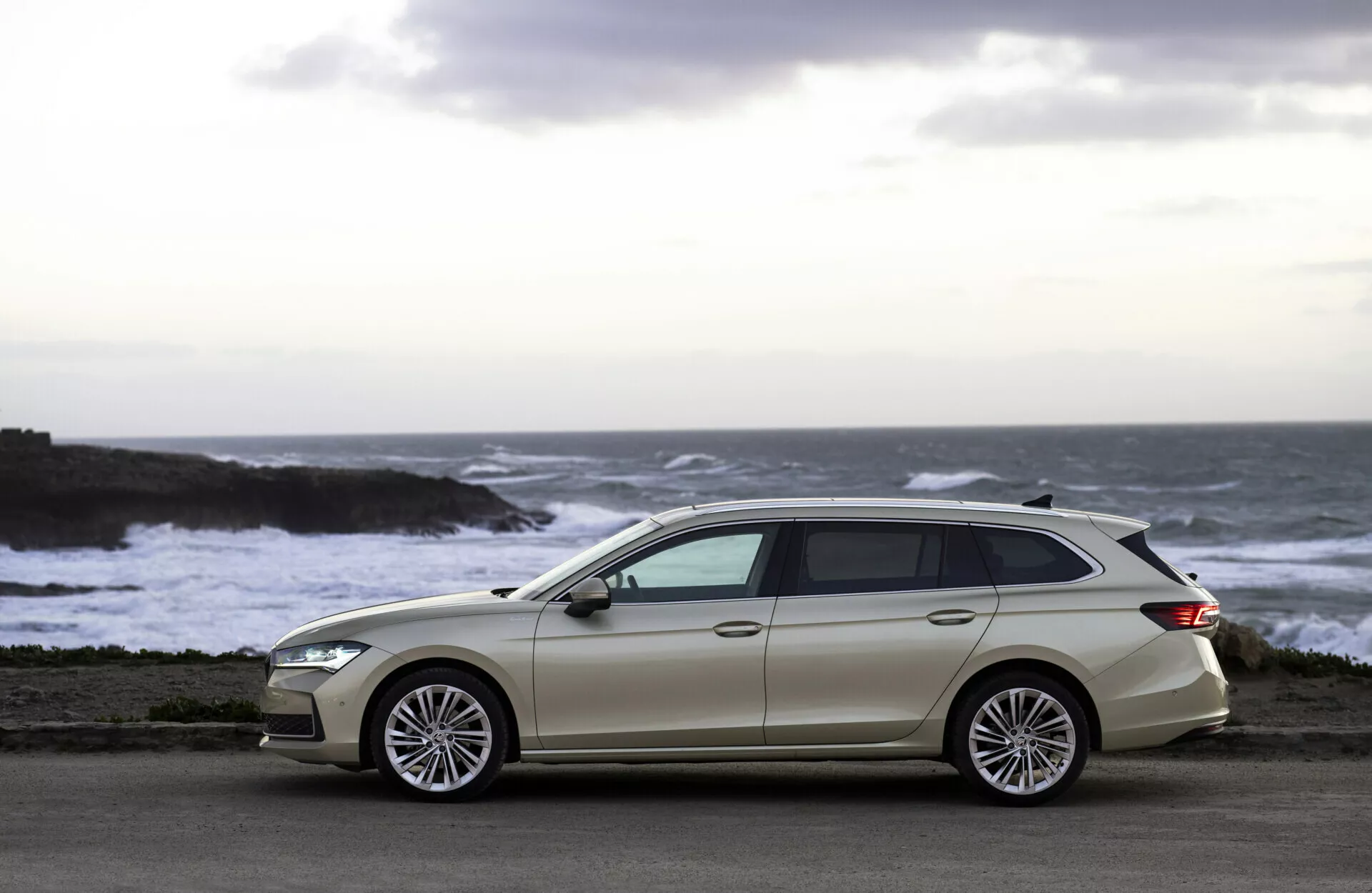 Ljusfärgad kombi Skoda Superb Combi 2024 parkerad på en kustväg med utsikt över havet, med en sluttande taklinje, fem dörrar, stora flerspakiga legeringshjul, kromfönstertrimning och karossfärgade sidospeglar mot en bakgrund av kraschande vågor och ett varmt skymningsljus.