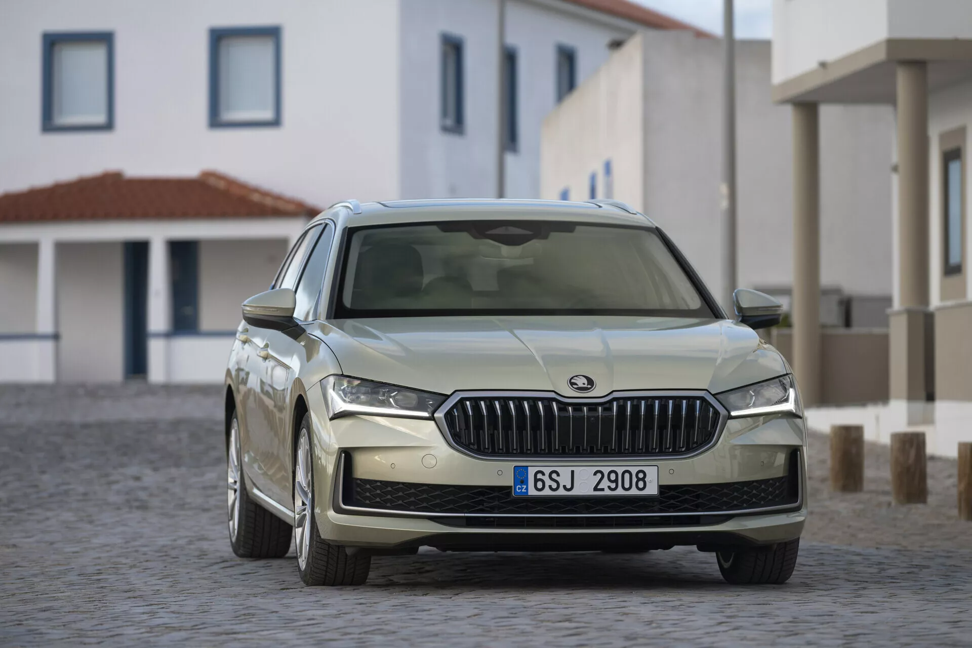 Sedan Skoda Superb Combi 2024 i dynamisk trekvartsutsikt som visar LED-strålkastare och sportiga legeringshjul på en kullerstensgata under varmt solljus