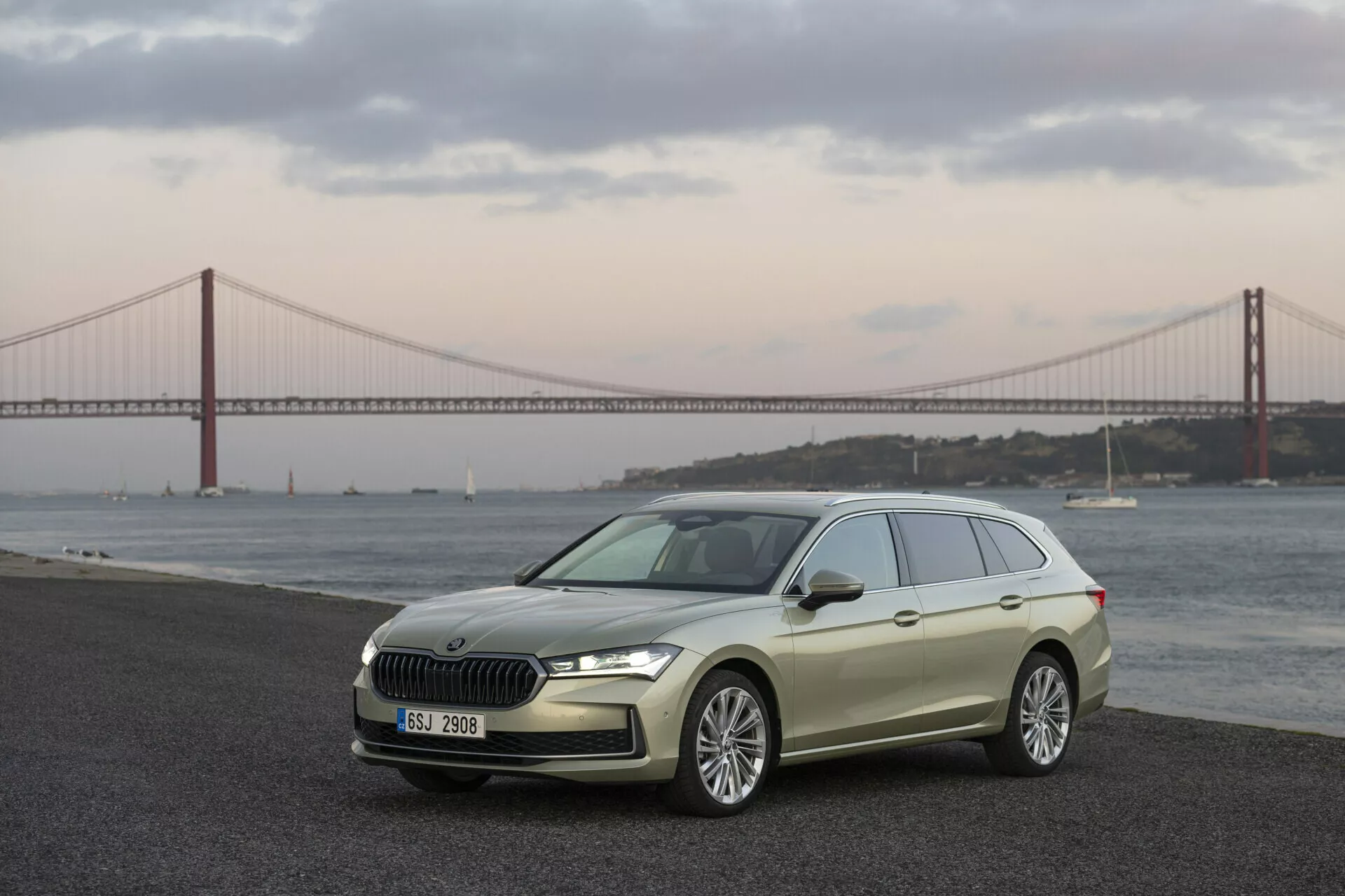 2024 Skoda Superb Combi framställd i en tre fjärdedels framvy som betonar den eleganta hatchbackdesignen, frontgrillen med kromfinish, LED-strålkastare och legeringshjul mot en naturskön bakgrund med en vattenkropp och bro i skymningen eller gryningen