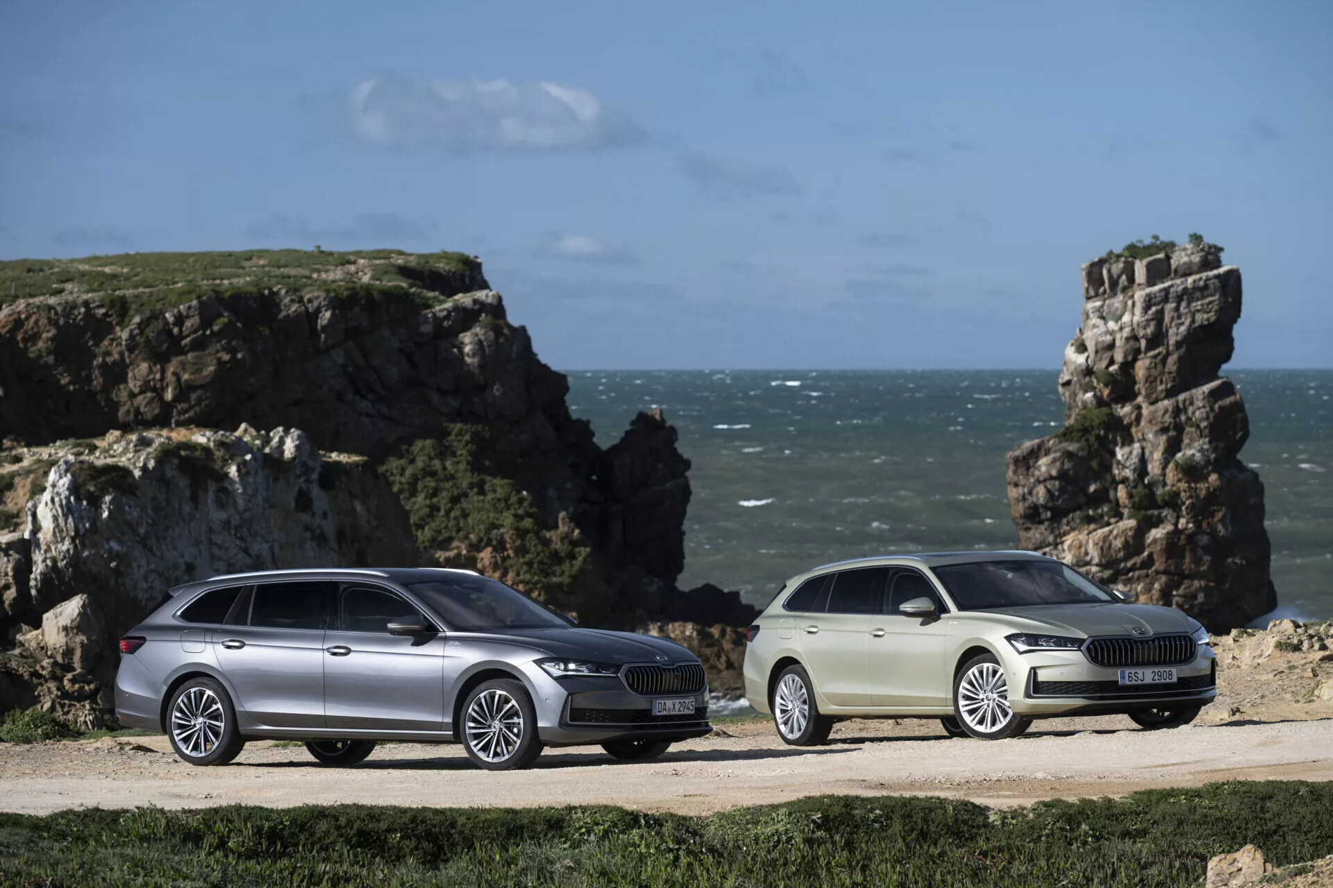 Skoda Superb Combi och sedan modeller från 2024 parkerade sida vid sida på en kustnära grusväg med en klippig kustklippa och en blå himmel i bakgrunden, med utökad utrymme för last, aerodynamisk design, LED-strålkastare och legeringshjul, vilket betonar deras äventyrliga och livsstilsinriktade tilltal.