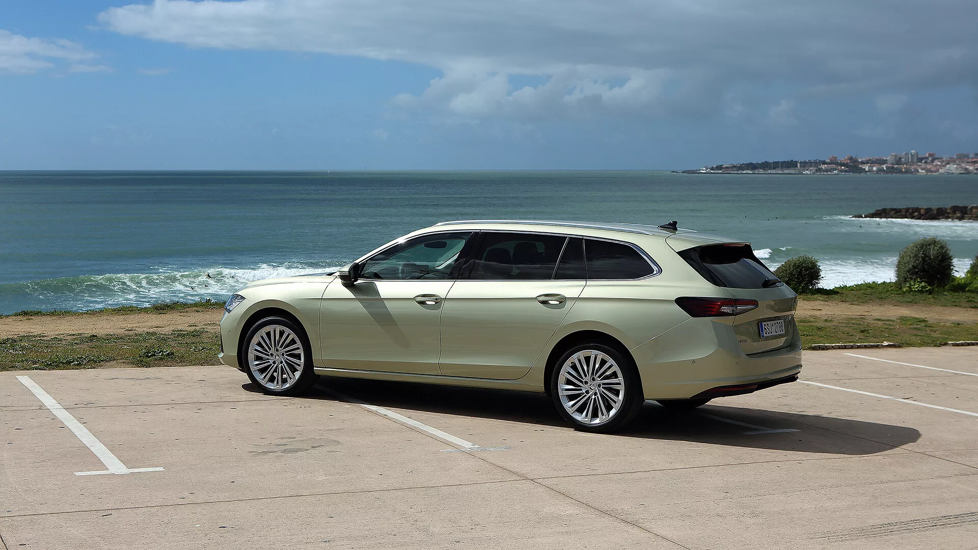 Skoda Superb Combi 2024 i vitt parkerad i vinkel på en parkeringsplats med utsikt över havet och kustlinjen under delvis molnig himmel, framhäver snygg design, modern strålkastare och grilldesign, stora lättmetallhjul och praktiska takräcken