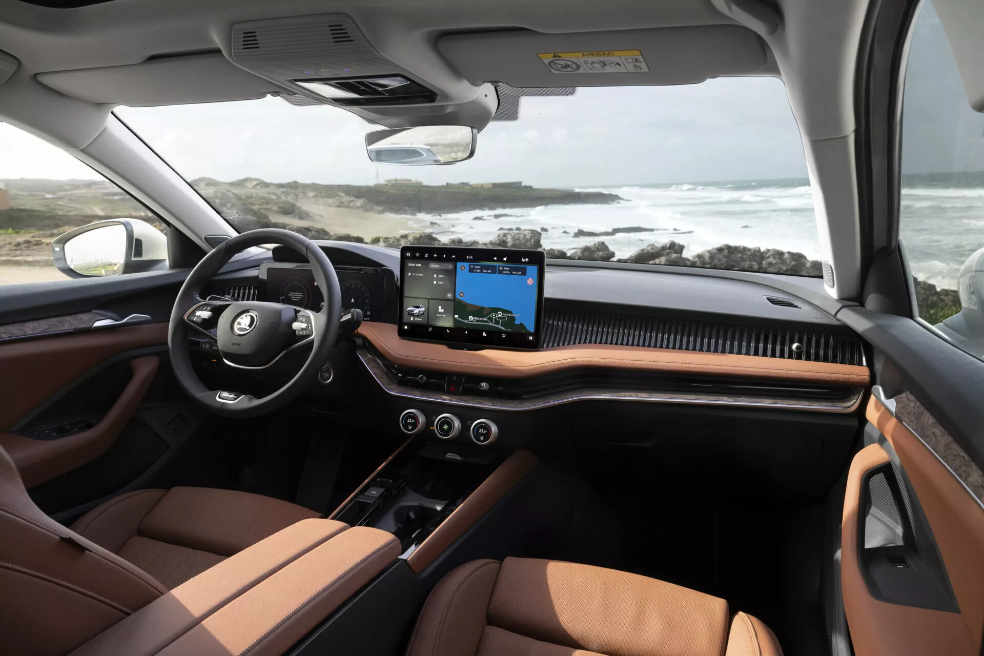 Interiörvy av Skoda Superb Combi 2024 som visar instrumentbräda och främre sittområde med tekniska funktioner och lyxig design, inklusive ett brett pekskärmsinfotainmentsystem, tvåfärgade lädersäten och ratt med monterade kontroller, mot bakgrund av en robust kustlinjescen genom vindrutan.