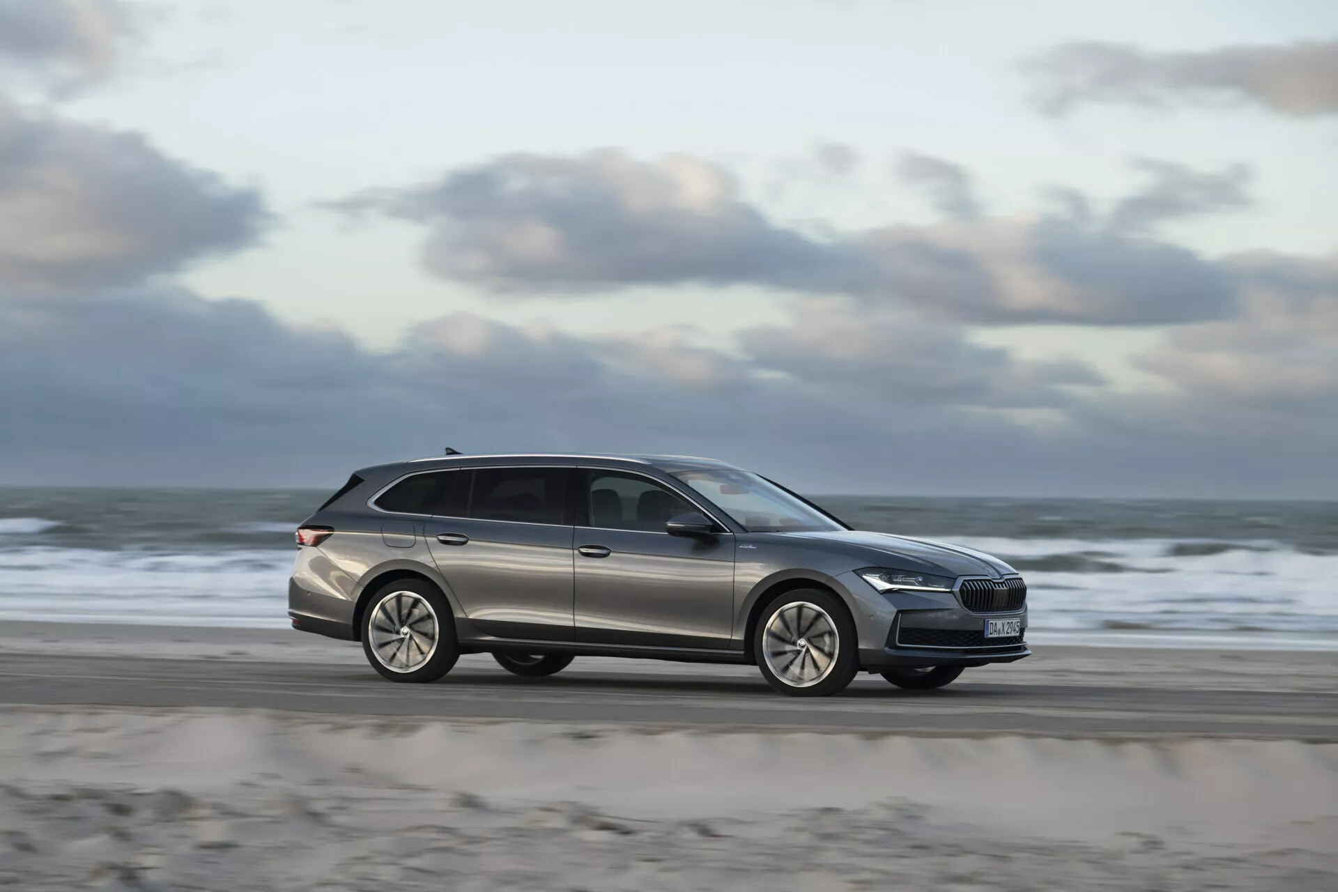 2024 Skoda Superb Combi kombi som kör längs en strandväg, visar modern design med sidoprofil, rörelseoskärpa indikerar hastighet, aluminiumhjul och takräcken för funktionalitet.