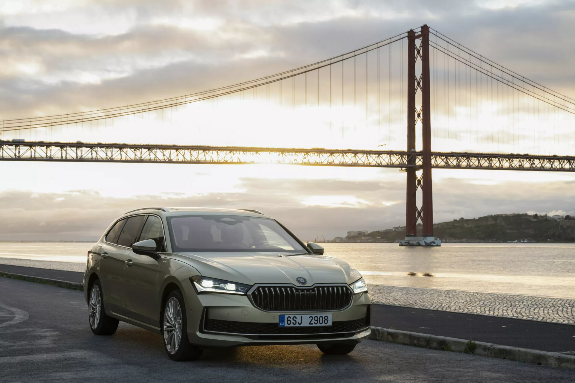 Lyxig SUV Skoda Superb Combi 2024 i gryningen, som visar moderna designfunktioner som vertikal frontgrill, snygga LED-strålkastare, konturerad huva och aerodynamisk profil nära en ikonisk bro.