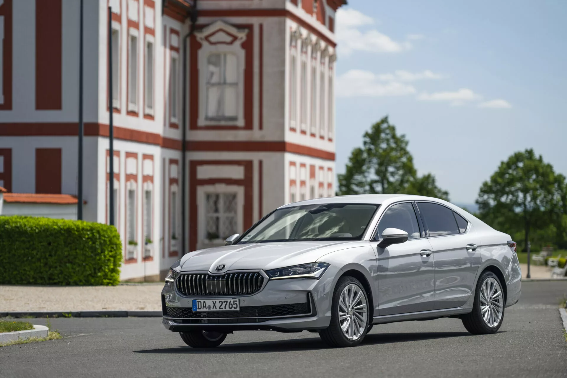 Lågvinkelbild av en silverfärgad Skoda Superb limousine 2024 parkerad på en stenlagd väg. Fotot framhäver bilens kromade frontgrill med vertikala spjälor, eleganta LED-strålkastare, dimljus och flerspokedaluminiumfälgar. Sidprofilen visar en ren design med subtila konturlinjer, kromade fönsterlister och inslag av avancerad LED-belysning och nyckelfri åtkomst. Den historiska byggnaden med röd och vit fasad i bakgrunden tillför djup till scenen, medan den klara blå himlen ger gott om naturligt ljus för att framhäva bilens egenskaper.