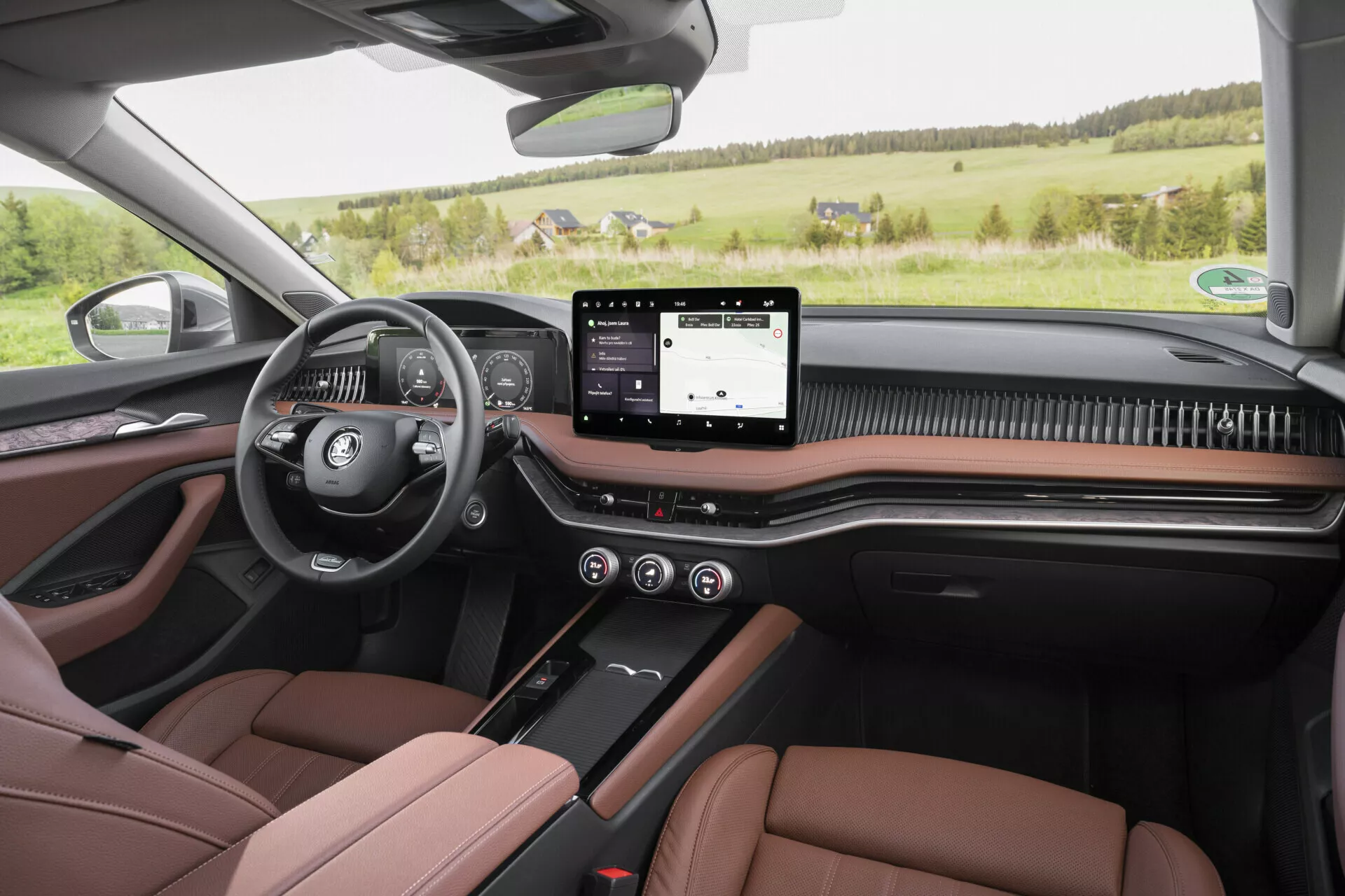En vy från första personens perspektiv från baksätet på 2024 Skoda Superb Limousine som visar en modern och lyxig interiör med en multifunktionell ratt på vänster sida, digital instrumentpanel, bred pekskärm för infotainmentsystemet, minimalistiska knappar för klimat- och säteskontroller under samt en kolfiberliknande trim. Bilden framhäver också tvåfärgade bruna och svarta lädersäten med kontrastsömmar, matchande dörrpaneler med elektriska kontroller, synliga högtalare för högkvalitativt ljudsystem och ett panoramasoltak, allt i en lantlig vy genom vindrutan.