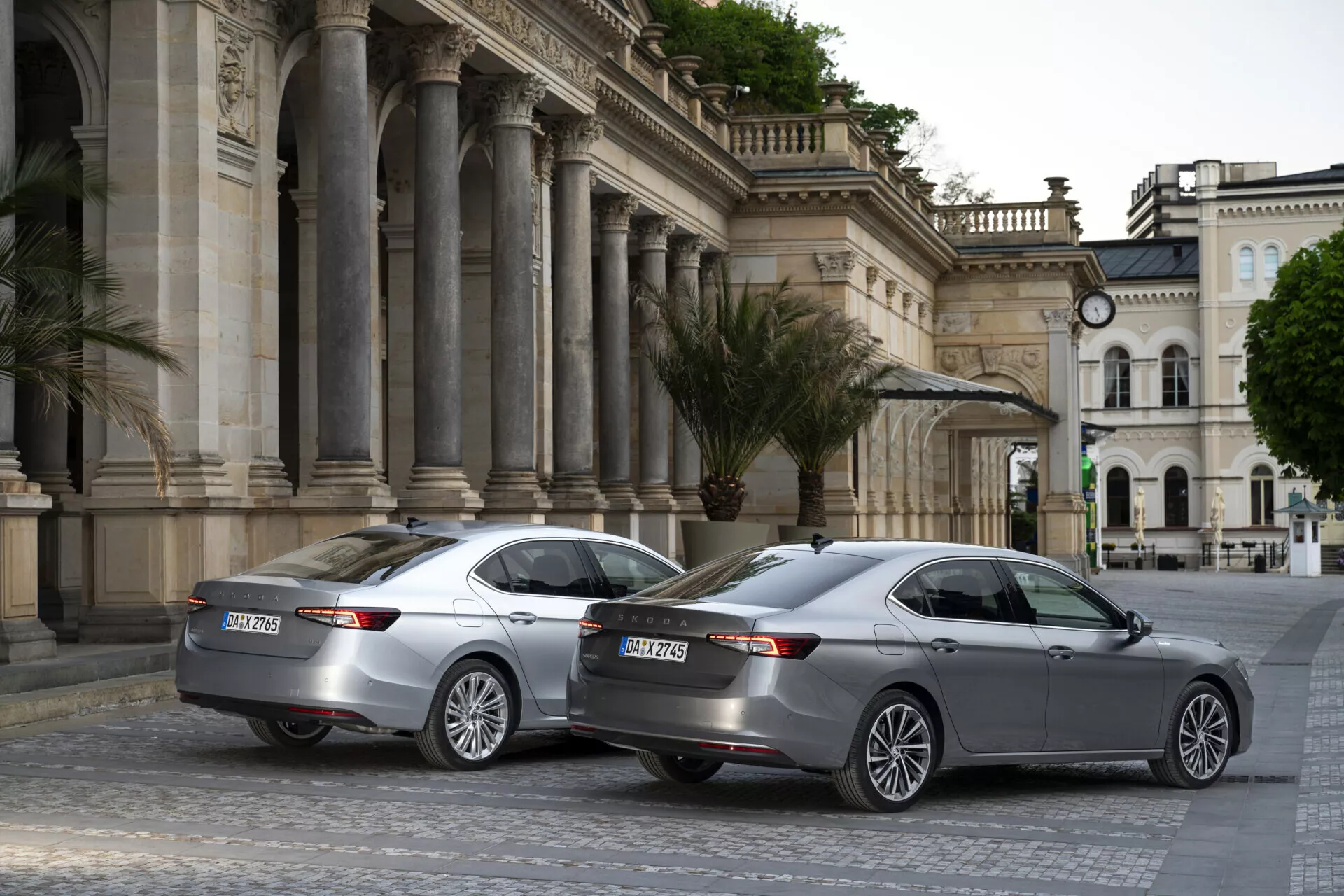 Bilden visar två identiska modeller av Skoda Superb 2024 sedans parkerade i ett urbant kulturcentrum med klassisk arkitektur. Fotot taget från en trekvarts vinkel bakifrån framhäver deras moderna och eleganta design med horisontella LED-bakljus, dubbla avgasrör, parkeringssensorer och flerspakiga lättmetallfälgar. Båda bilarna har hajfensantenner för avancerad uppkoppling och ett tredje bromsljus på den nedre delen av bakrutorna. Scenen är fångad i molnigt eller tidigt morgon/kvällsljus, vilket ger ett enhetligt och något dämpat ton till fotografiet.