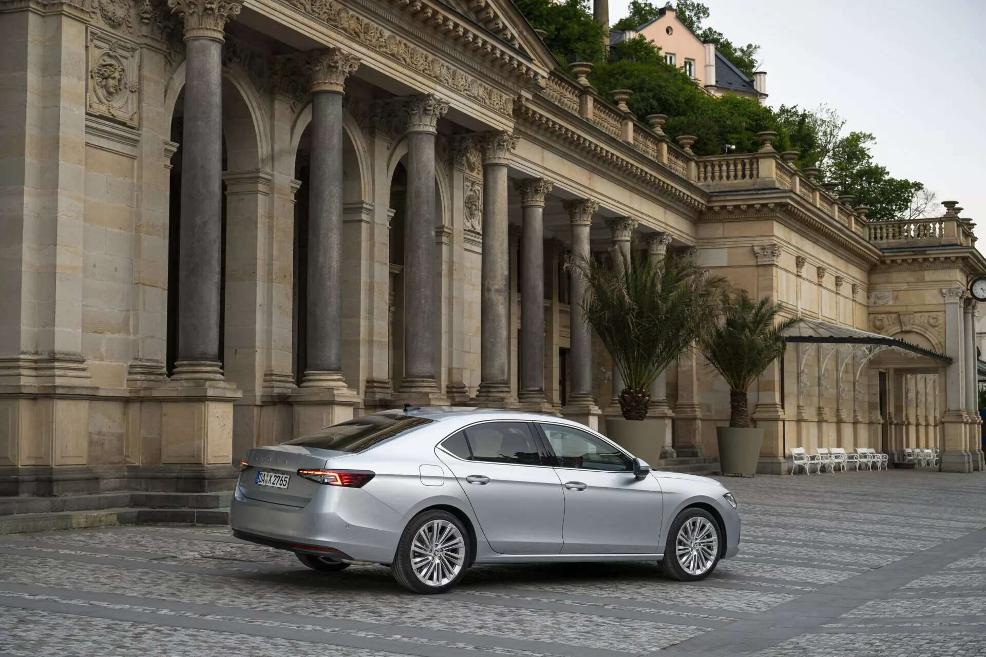 En omfattande vy av en silverfärgad Skoda Superb Limousine 2024 sedan, parkerad diagonalt på ett kullerstensbelagt torg med klassisk, utsmyckad arkitektur i bakgrunden. Bilden är tagen från en bakre trekvarts vinkel och visar de eleganta baklysena, bagageluckan, bakre stötfångaren och sidokonturlinjerna på fordonet. Den mjuka belysningen och lugna atmosfären antyder en mulen himmel eller gyllene timmen, och den stillastående bilen, med lättmetallfälgar och flerspårsdesign, är omgiven av gott om utrymme, vilket gör den till ett lämpligt motiv för bilkategorier, lyxsedaner eller arkitektur.