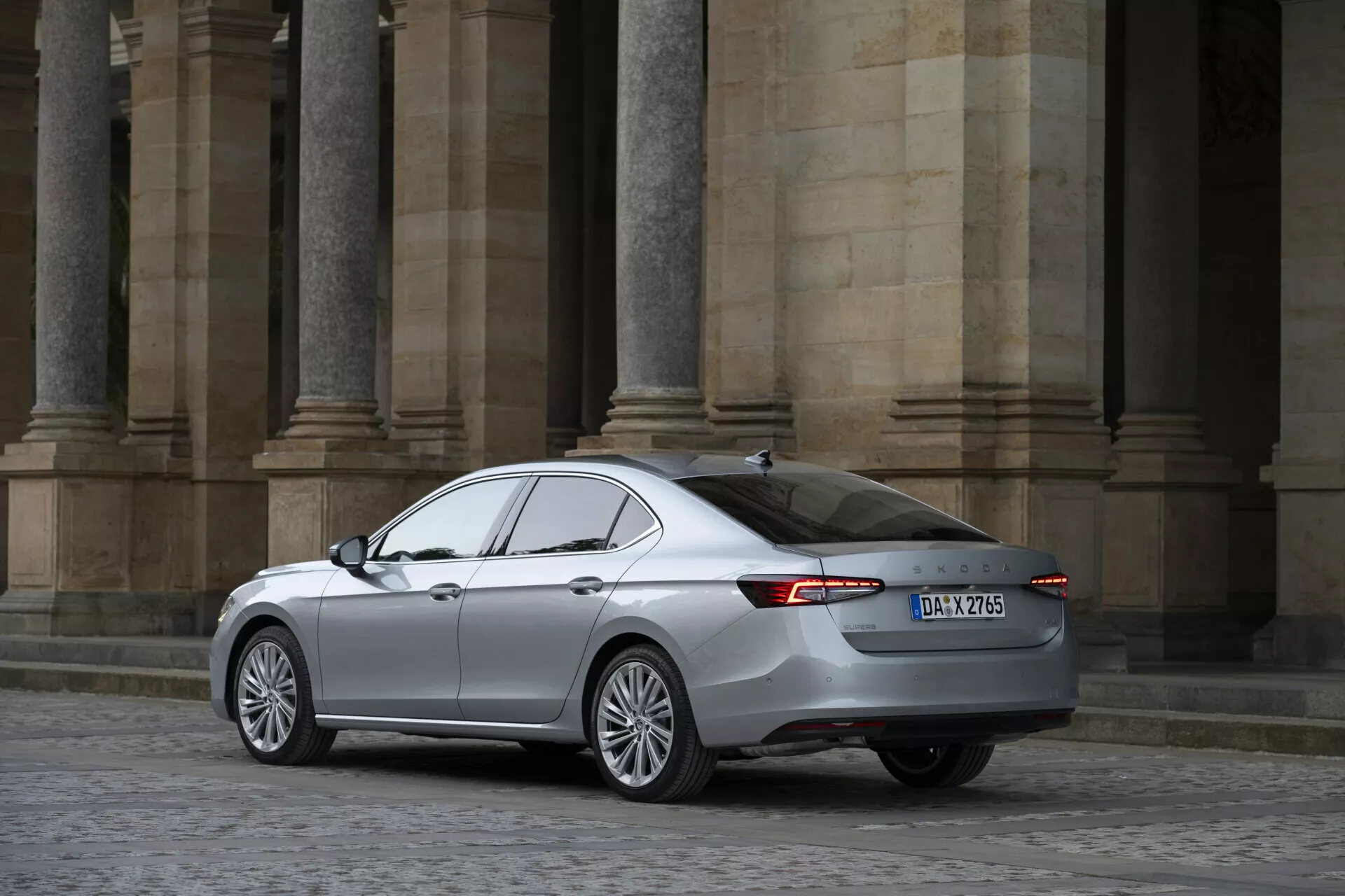 Bild av 2024 Skoda Superb sedan från en trekvarts bakvinkel. Med betoning på höger sida och bakdel har bilen lättmetallfälgar, LED-bakljus, strömlinjeformad kaross med metalliclack, tonade fönster och synliga emblem. Bilden, tagen på medelavstånd, visar fordonet utan hinder med klassisk arkitektur i bakgrunden.