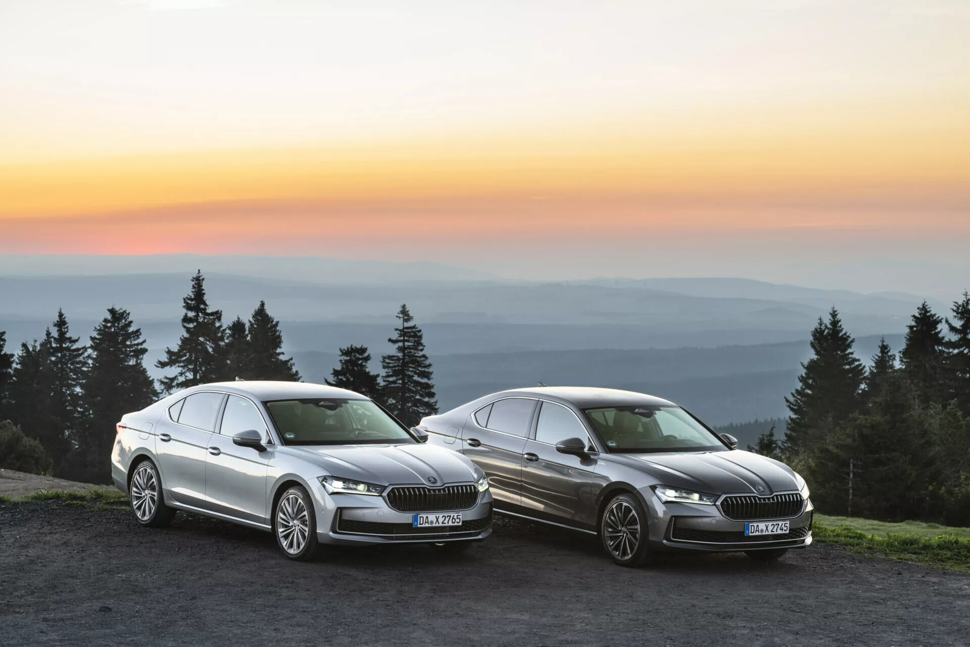En bild på två Skoda Superb 2024-bilar placerade på en upphöjd kulle vid solnedgång med ett vidsträckt landskap i bakgrunden. Båda bilarna är vinklade mot kameran i ungefär 45 grader, visar sina grillar, strålkastare, vindrutor och eleganta karossdesigner. Ljuset från guldtimmen framhäver bilarnas metalliska ytor och lyxiga detaljer, betonar deras moderna funktioner och avancerade LED-strålkastare. Bilarna är placerade måttligt nära kameran, upptar största delen av ramen med en lätt överlappning och balanserad komposition.