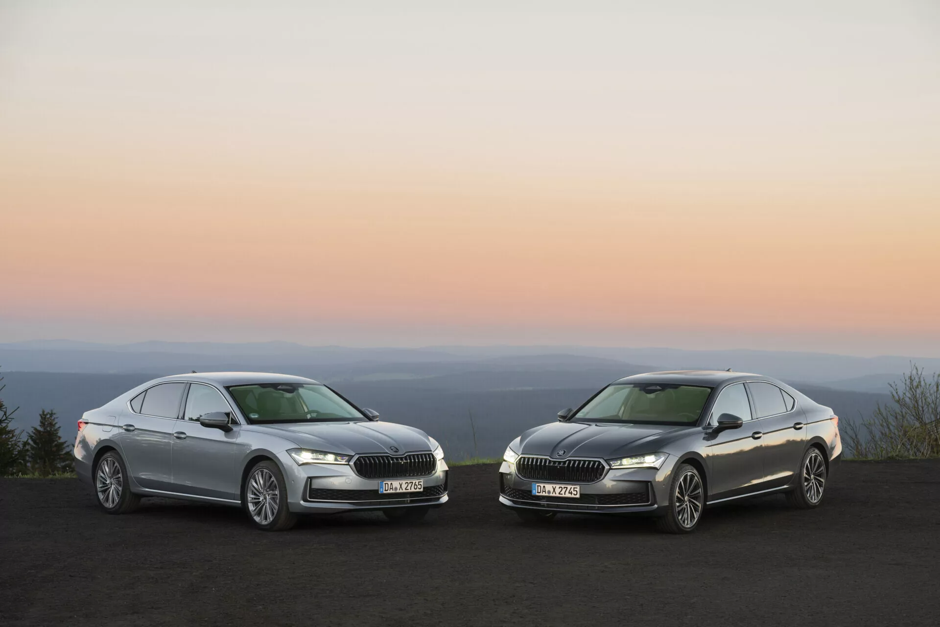 Högvinklad fotografi av två Skoda Superb 2024-sedaner parkerade bredvid varandra vid soluppgång eller solnedgång, som visar bilarnas framsida, inklusive huvar, strålkastare och grill med kromdetaljer. Lyxbilarna med avancerad belysningsteknik är markerade av naturligt omgivande ljus, vilket betonar bilarnas design, varumärke och strömlinjeformade karosseri mot en mjuk orange-rosa gradienthimmel.