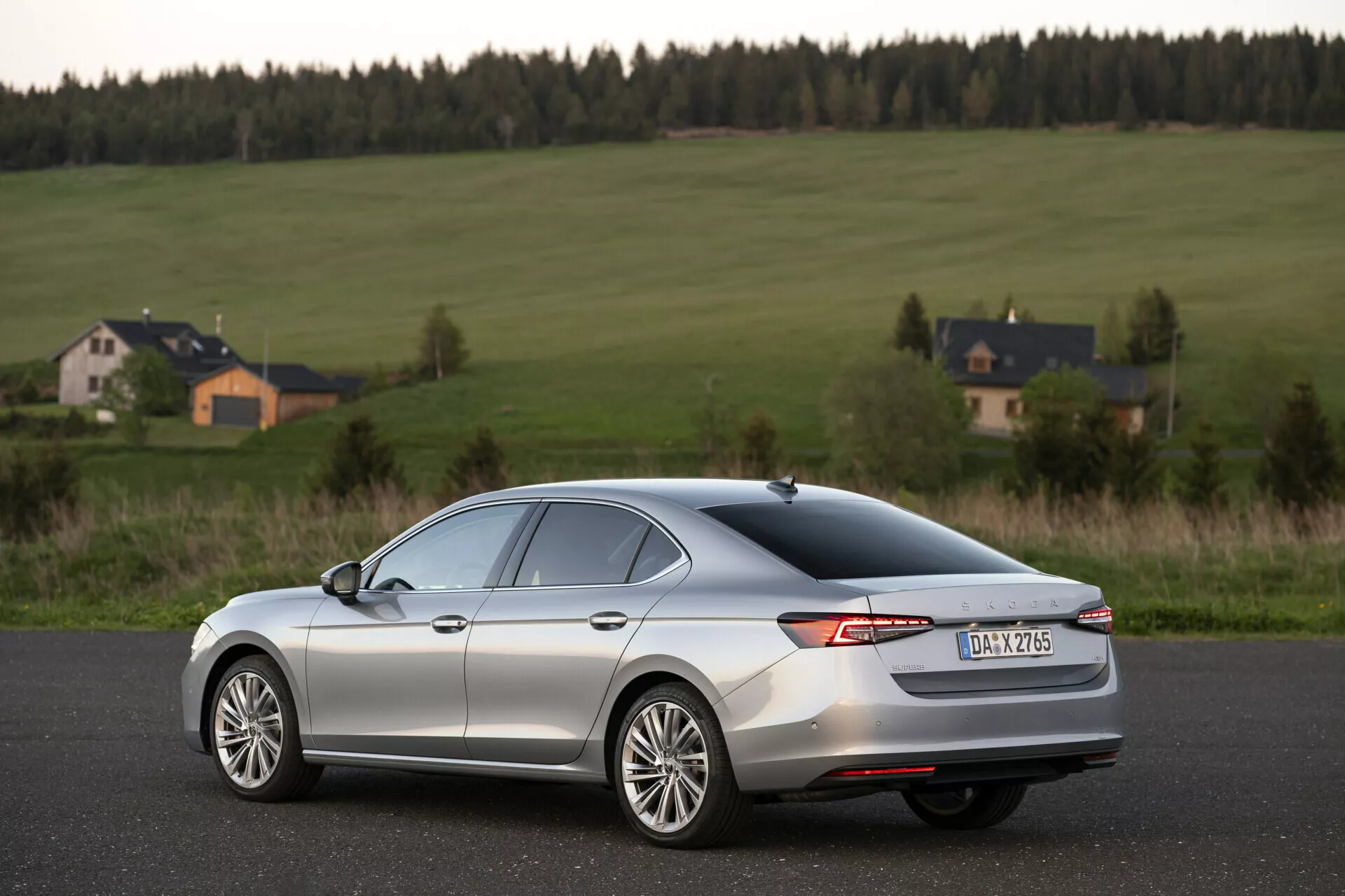 Bilden visar en Skoda Superb Limousine 2024, en modern sedan med en slank, aerodynamisk design och metallisk silverlack, fångad från en bakre trekvarts vinkel. Bilen står stilla på en slät asfalterad väg i en lantlig miljö med öppna fält och hus i bakgrunden. Kännetecken inkluderar omslutande LED-baklyktor, en bakre bagagespoiler, flerspokes fälgar, en avimningsraster på bakrutan och en hajfensantenn på taket. Fotot, taget under den gyllene timmen, har ett grunt skärpedjup fokuserat på bilen, med ett naturligt ljus som framhäver dess konturer.