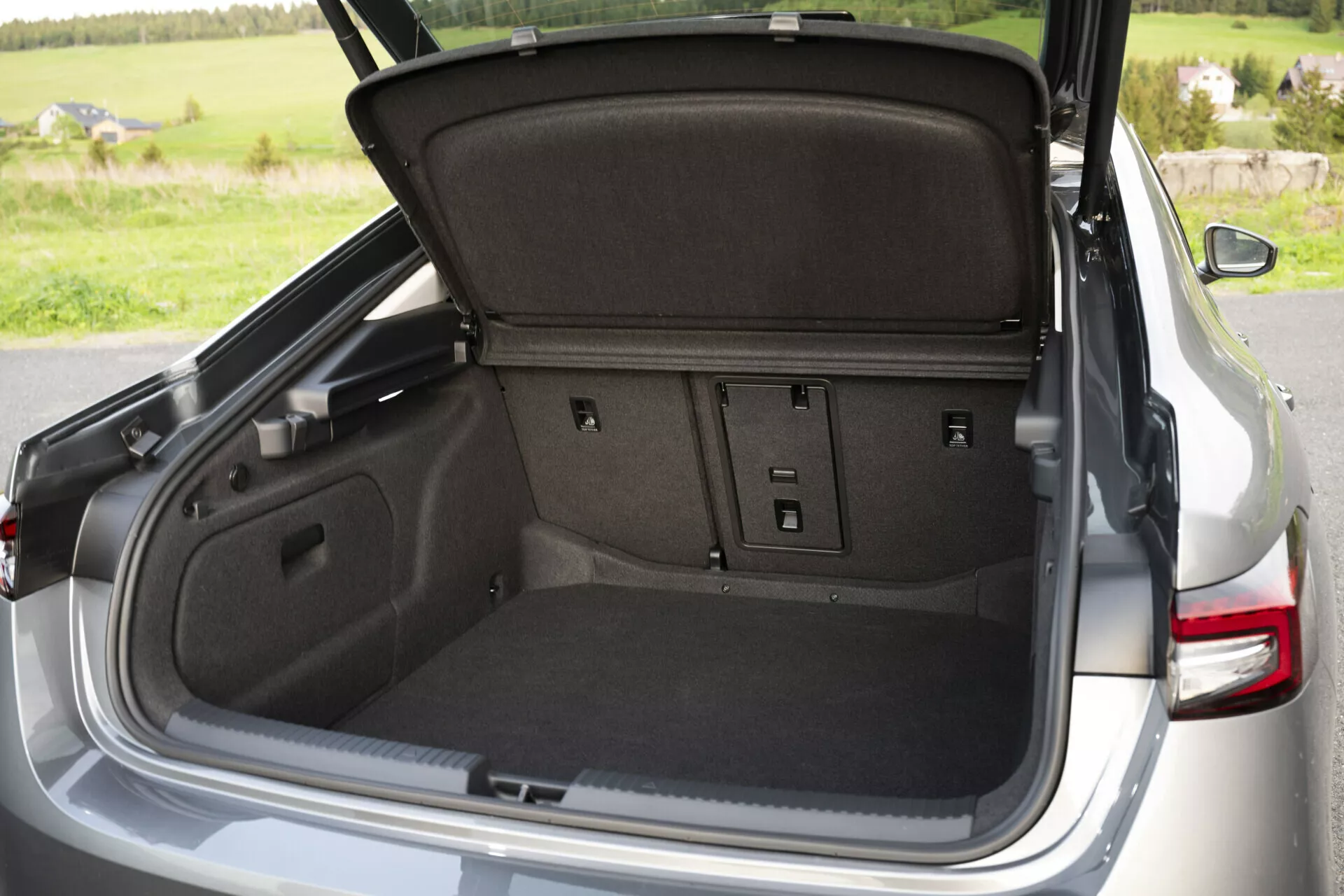 Närbild av den öppna bagageutrymmet på Skoda Superb 2024 sedan, som visar en rymlig matta-fodrad lastutrymme med delbara baksäten, lastkrokar, bagagebelysning, åtkomstpaneler, strömuttag och ett utdragbart insynsskydd, mot en bakgrund av naturliga utomhusomgivningar.